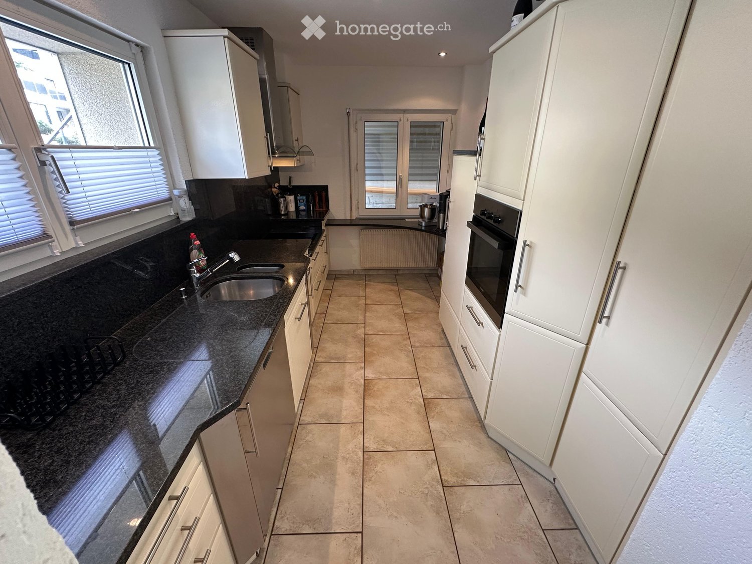 Modern kitchen with white cabinets, dark granite countertops, stainless steel sink, and built-in oven. Features large windows with blinds and tiled flooring.
