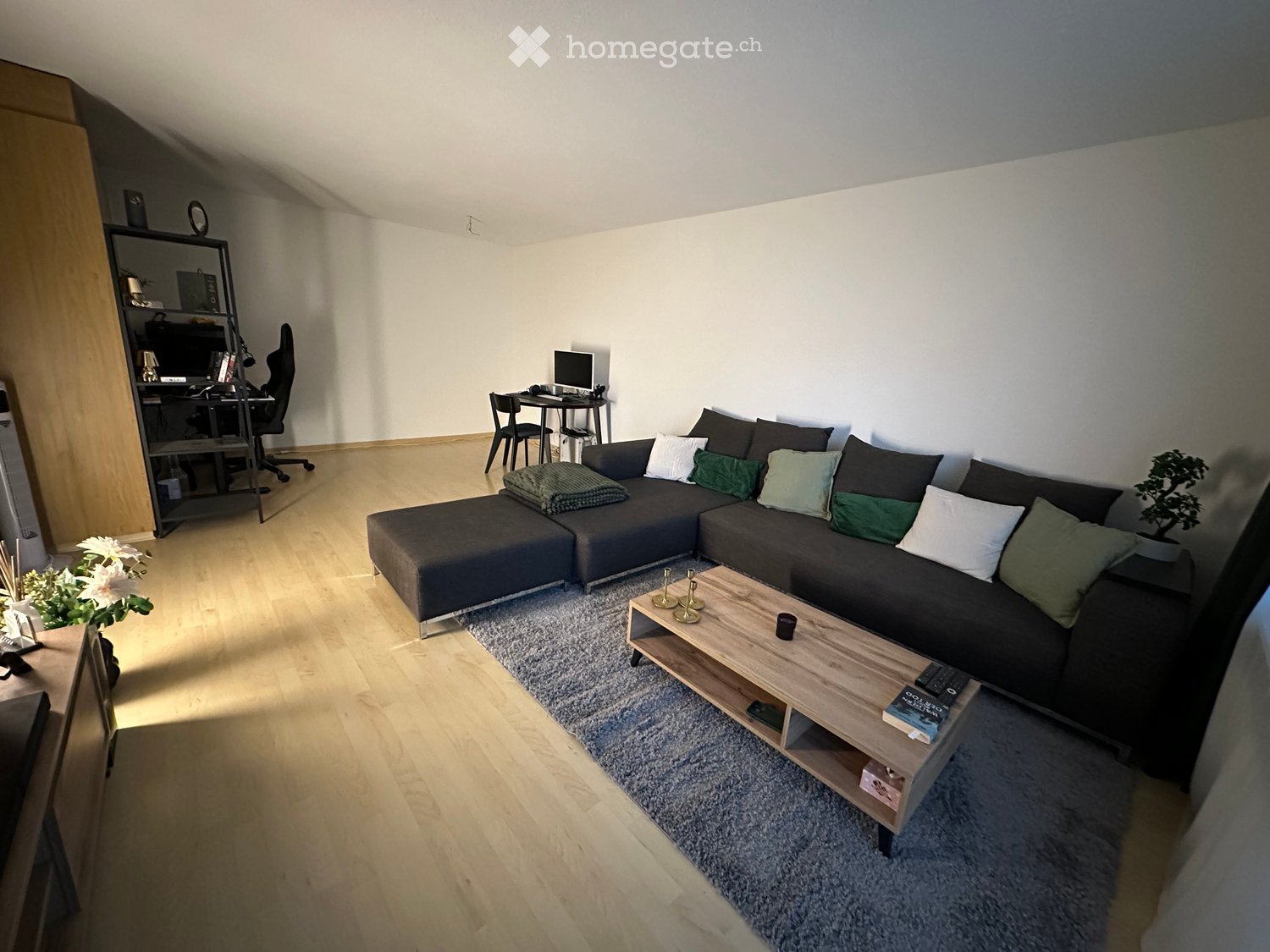 Modern living room with a dark grey sectional sofa, wooden coffee table, small dining area with a table and chairs, and a bookshelf with a laptop and decorative items.