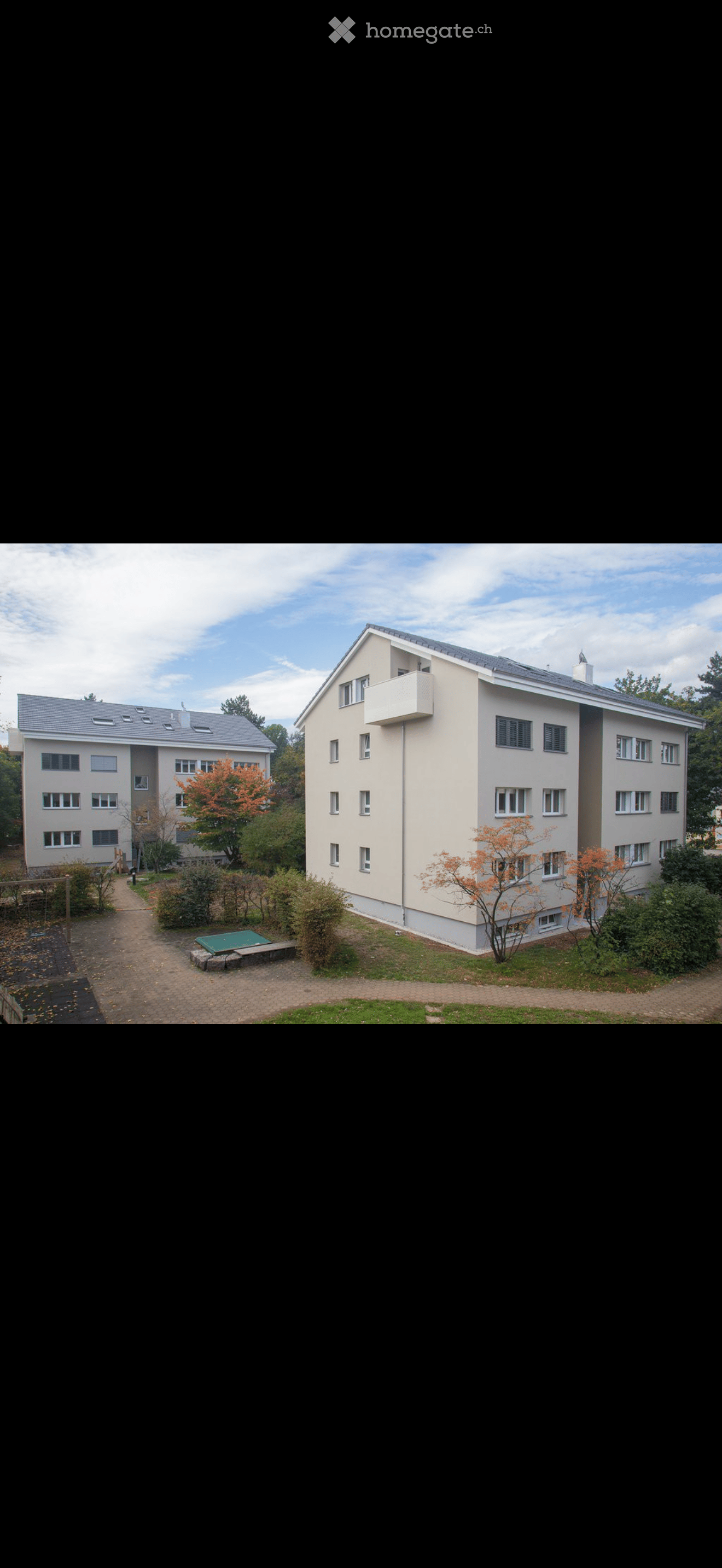 Two-story residential building with white walls, multiple windows, and a small balcony on the right side. The building is surrounded by greenery and a paved pathway.