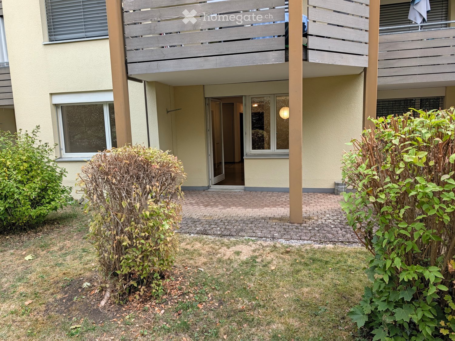 Garden area with a paved pathway leading to a building entrance, surrounded by shrubs and grass.