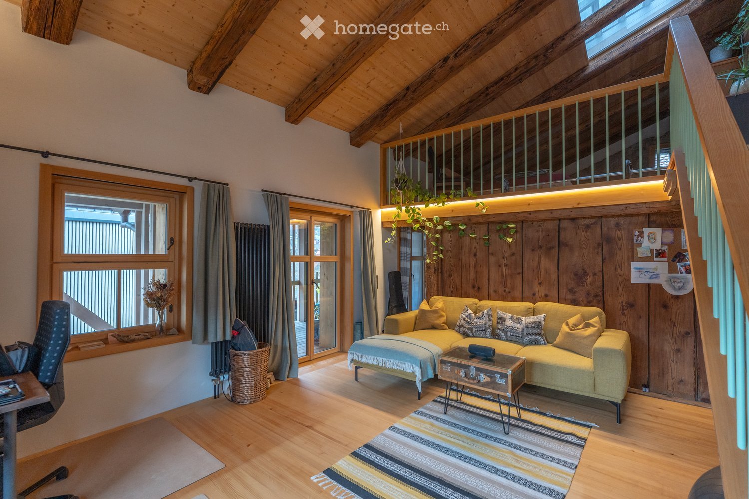 Cozy living room with exposed wooden beams, large windows, and a balcony. Features a light yellow sectional sofa, coffee table, wicker basket, and a hanging plant.