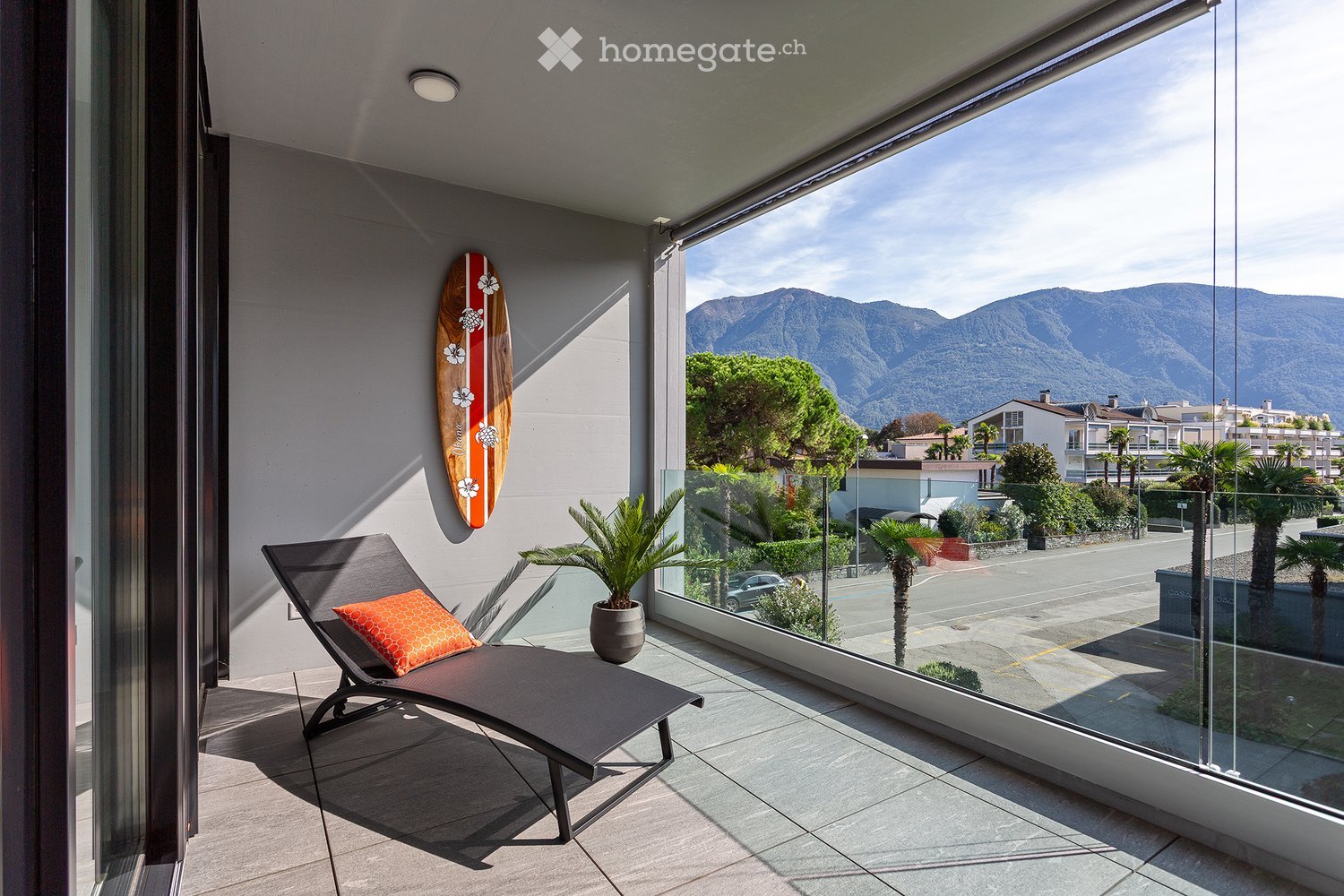 Balcony with a lounge chair, potted plant, and surfboard on the wall, offering a scenic mountain view.