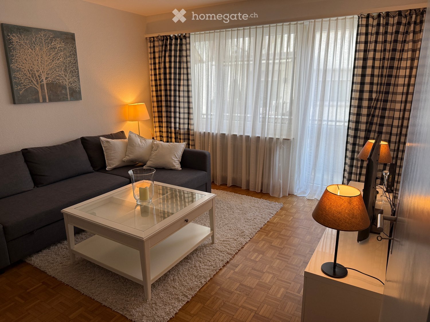 Modern living room with a dark grey sofa, white coffee table, two table lamps, a floor lamp, and a television on a stand. The room features large windows with checkered curtains and a white rug on the wooden floor.