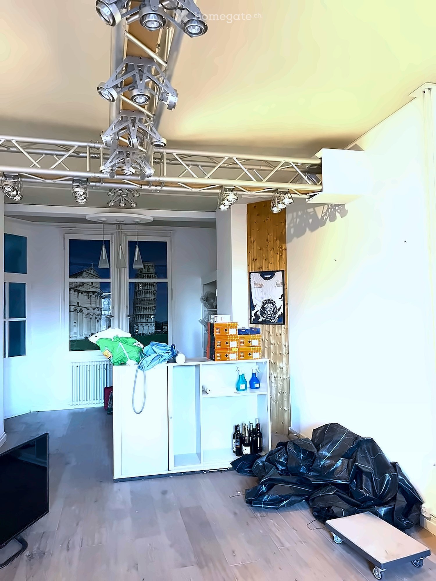 The room features a modern, industrial design with exposed lighting rigs, a large window showing a scenic view, a cabinet with bottles, and a covered object on the floor.