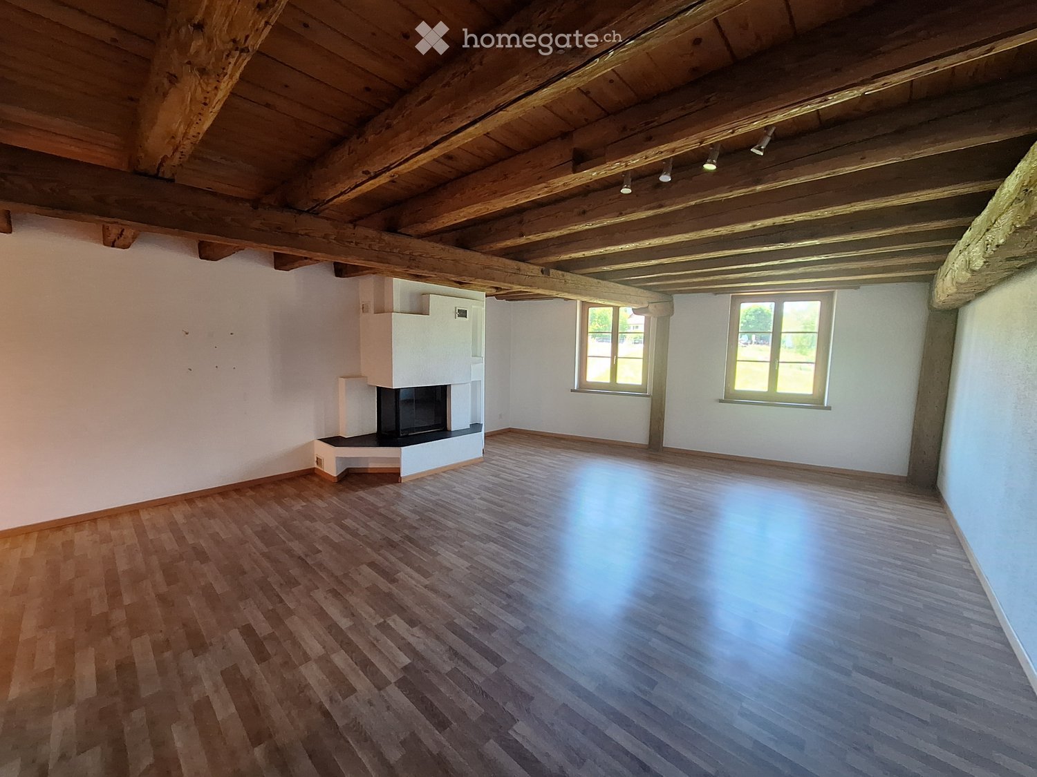 Empty room with wooden floor, white walls, exposed wooden ceiling beams, and a fireplace with built-in shelving.