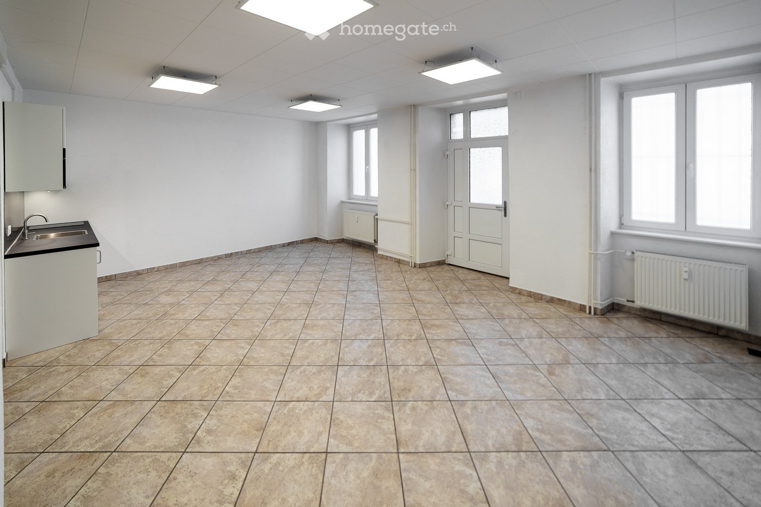 Empty room with tiled floor, white walls, and a small sink area. The room has several windows and a door leading to another space.