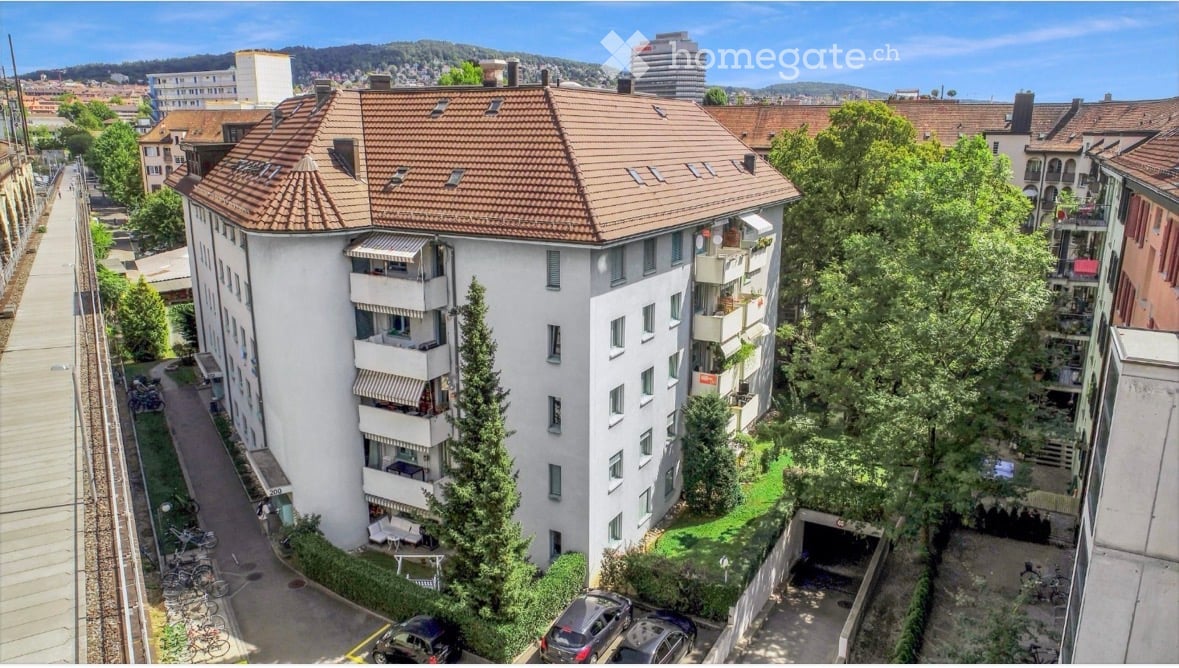 Multi-story residential building with a red-tiled roof, balconies, and surrounding greenery. Includes a parking area and garage.