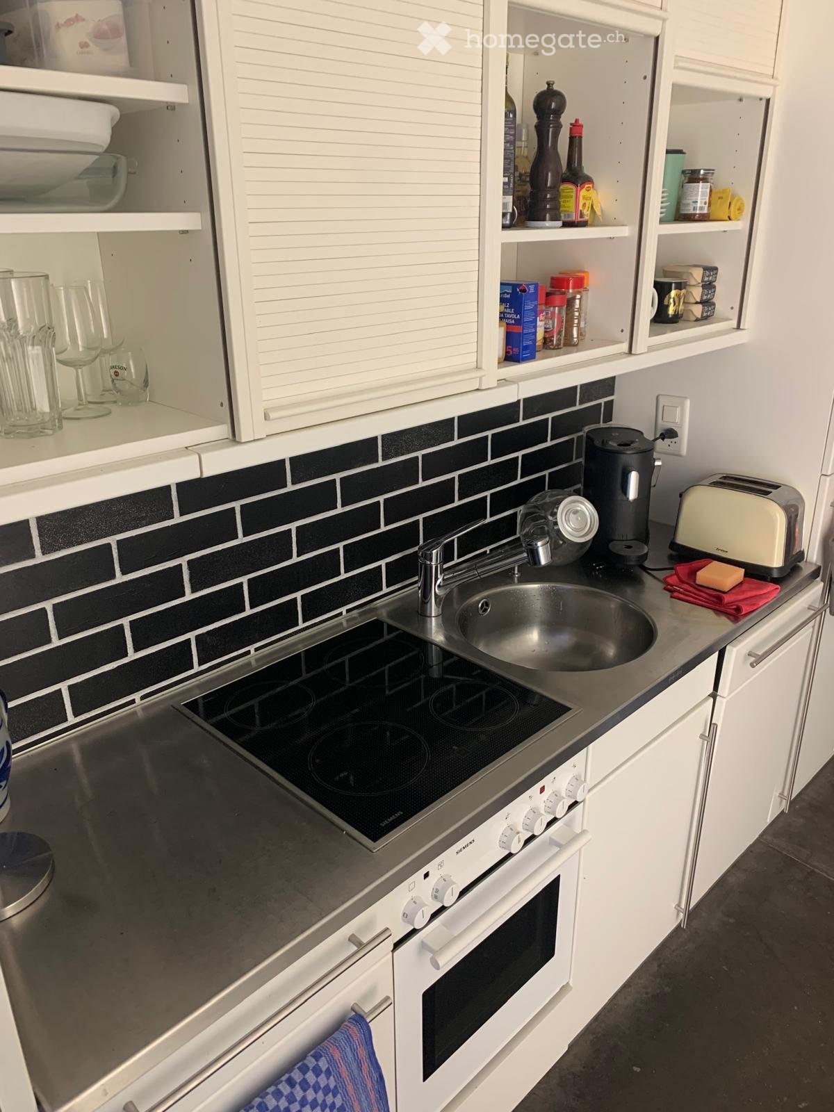 Modern kitchen with white cabinets, stainless steel sink, induction cooktop, oven, coffee maker, and toaster. Shelves hold various kitchen items including bottles, cups, and food containers.