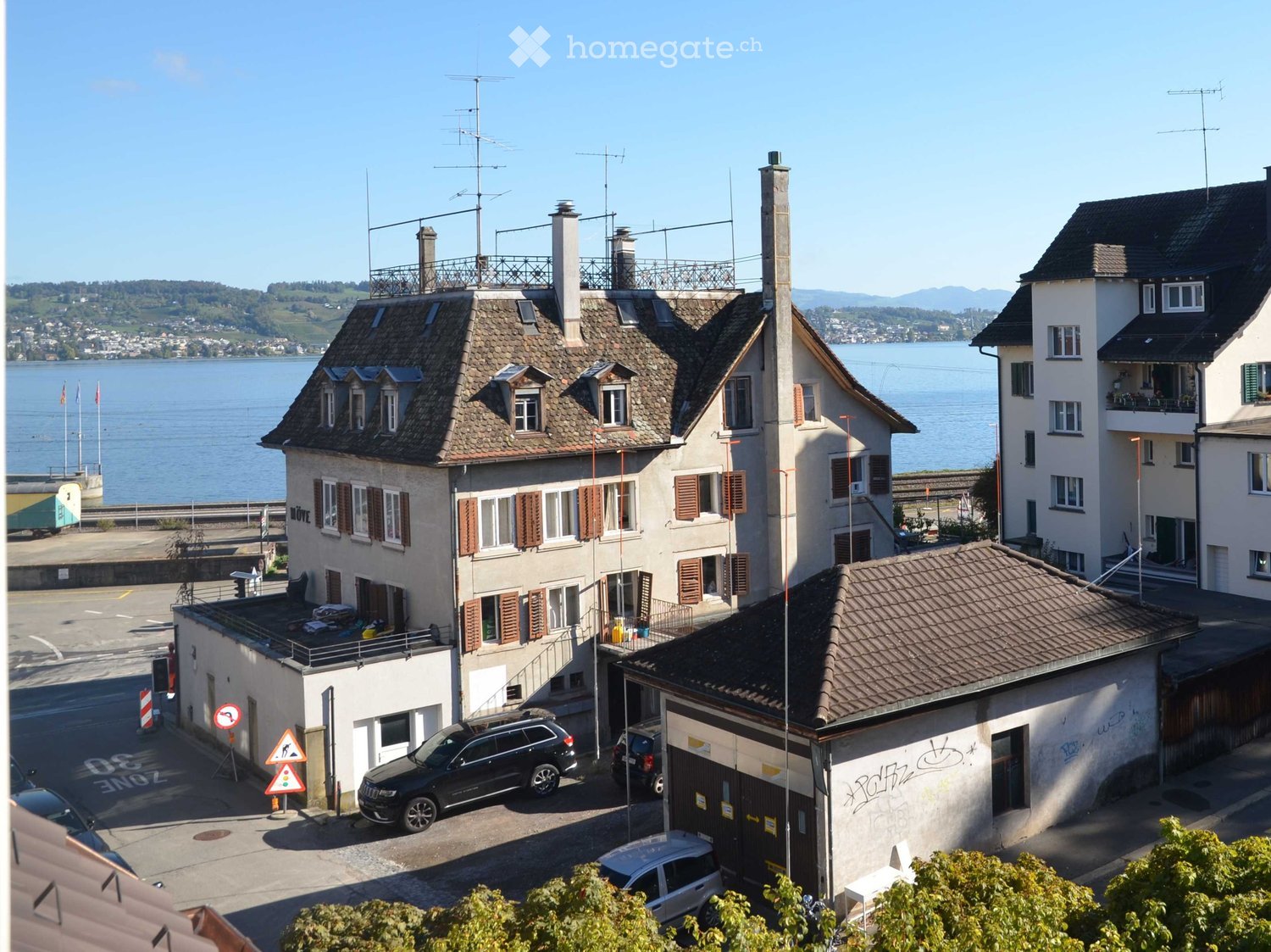 Multi-story residential building with a tiled roof, chimneys, and multiple windows, situated near a waterfront with visible lake and distant mountains.