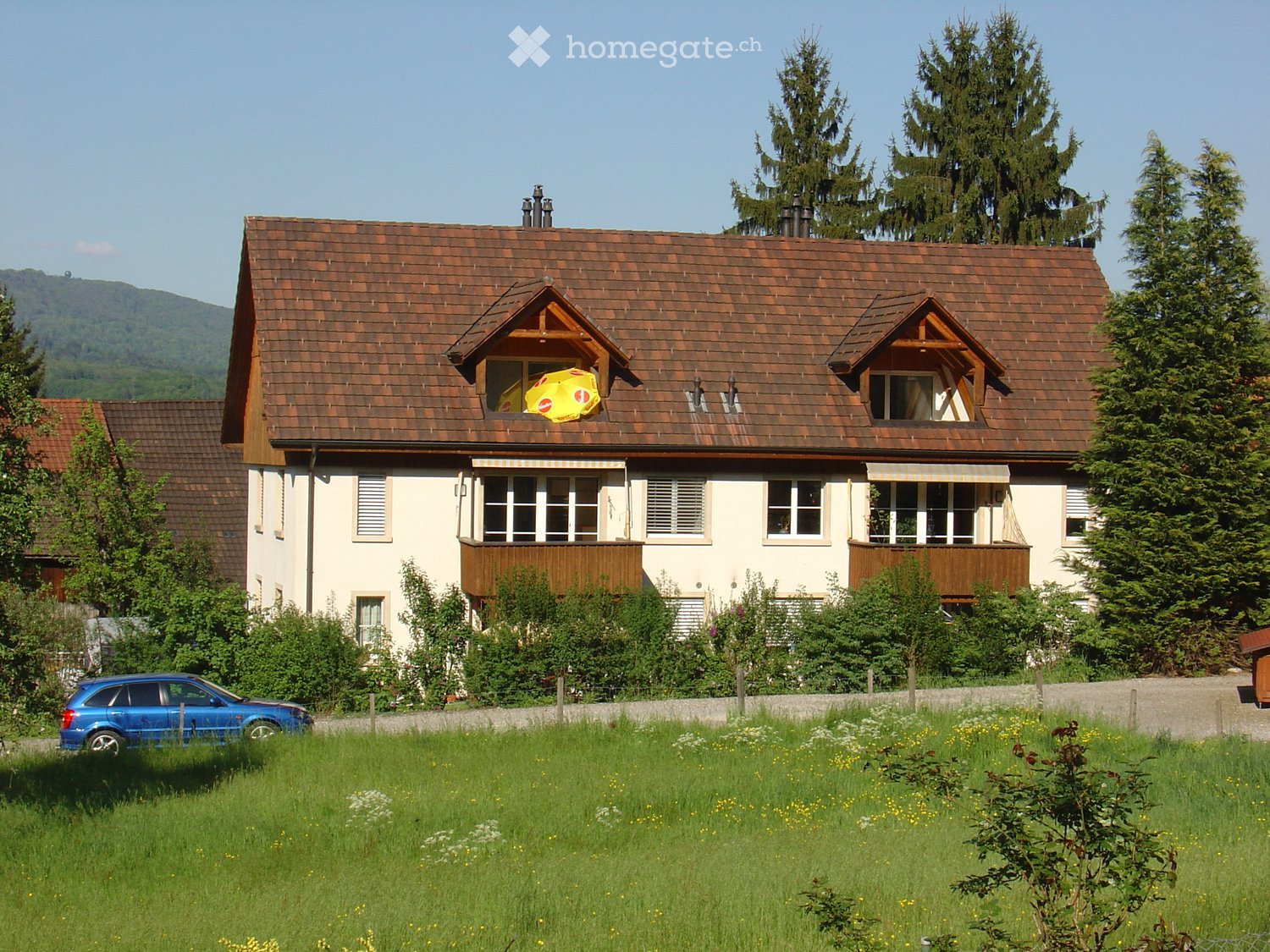Two-story house with a brown tiled roof, white walls, multiple windows, and a small balcony. A blue car is parked in front, and the house is surrounded by a well-maintained lawn and trees.