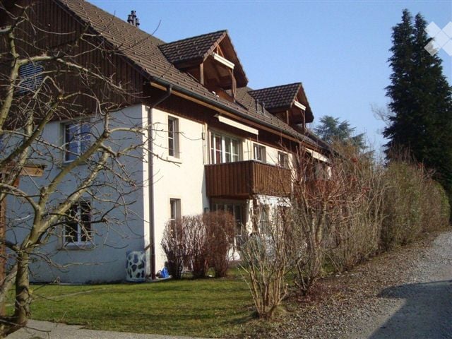 A two-story house with white walls, dark wooden roof, and a balcony on the upper floor. The house is surrounded by a well-maintained lawn, shrubs, and trees.