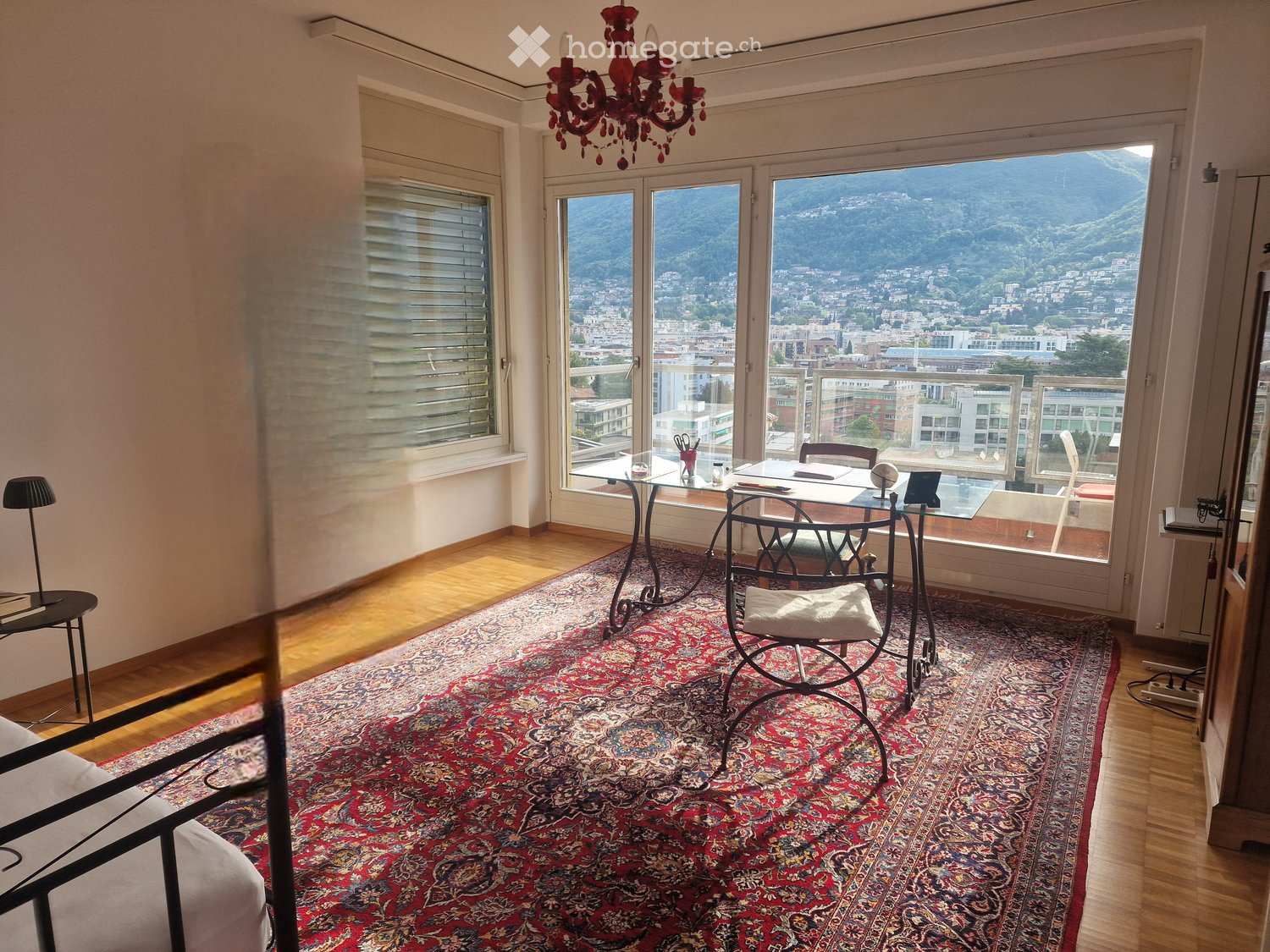 Bright room with large windows offering a scenic mountain view, furnished with a red patterned rug, a glass table, chairs, a chandelier, and a lamp.