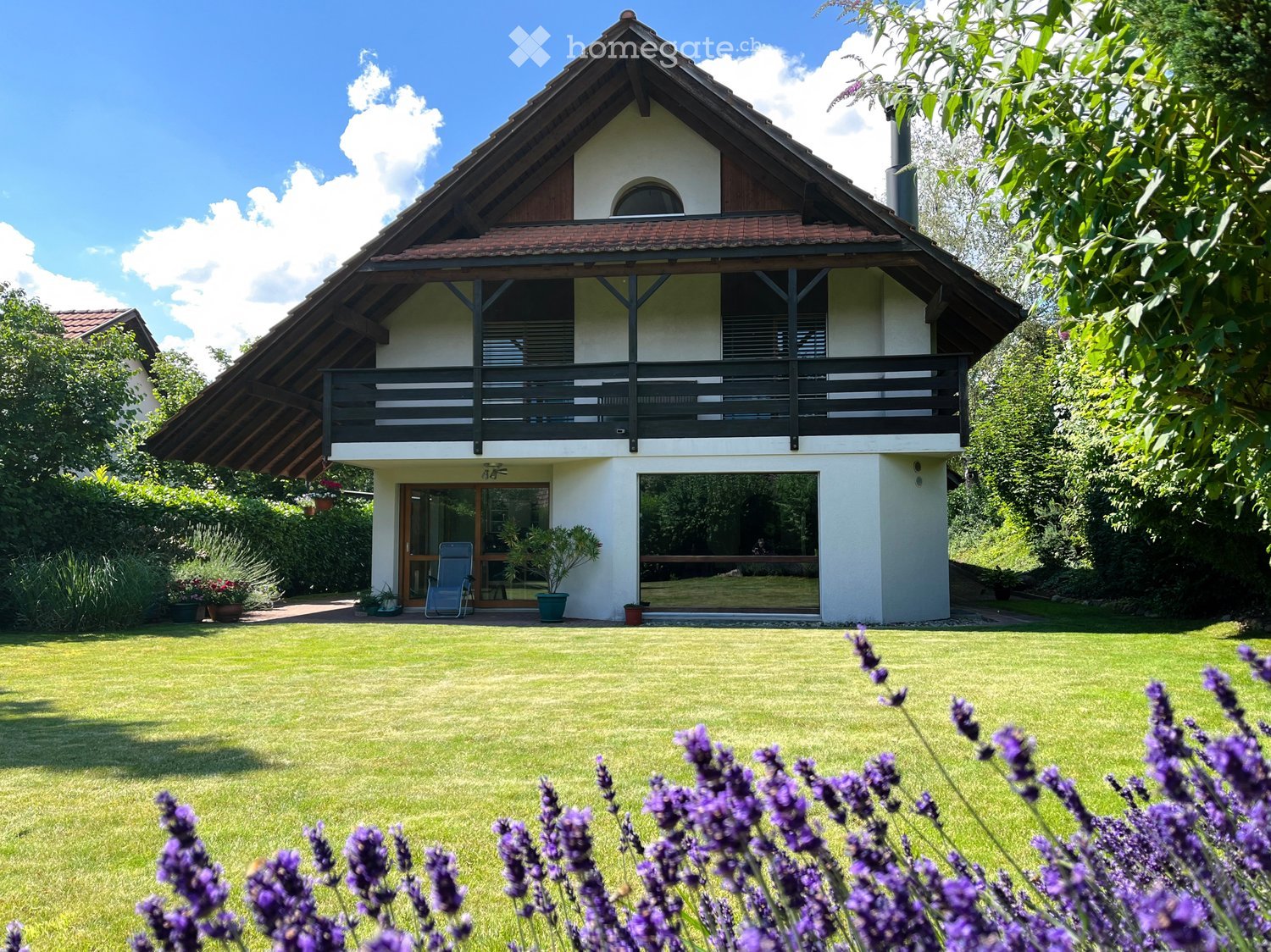 Two-story house with a balcony, surrounded by a well-maintained lawn and garden with lavender plants.