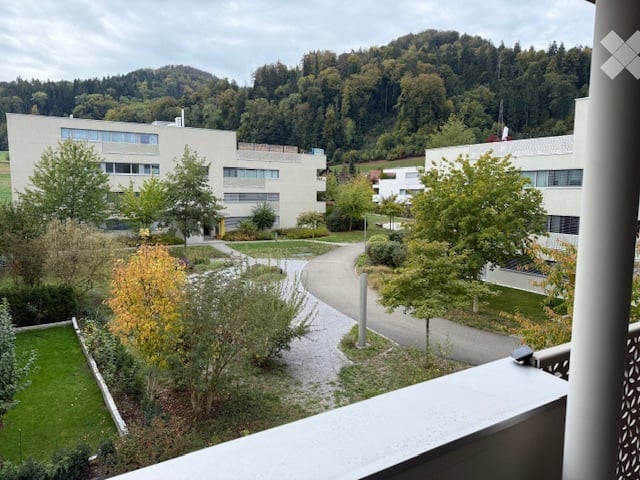 Balcony overlooking a landscaped courtyard with modern buildings, trees, and a distant forested hill.
