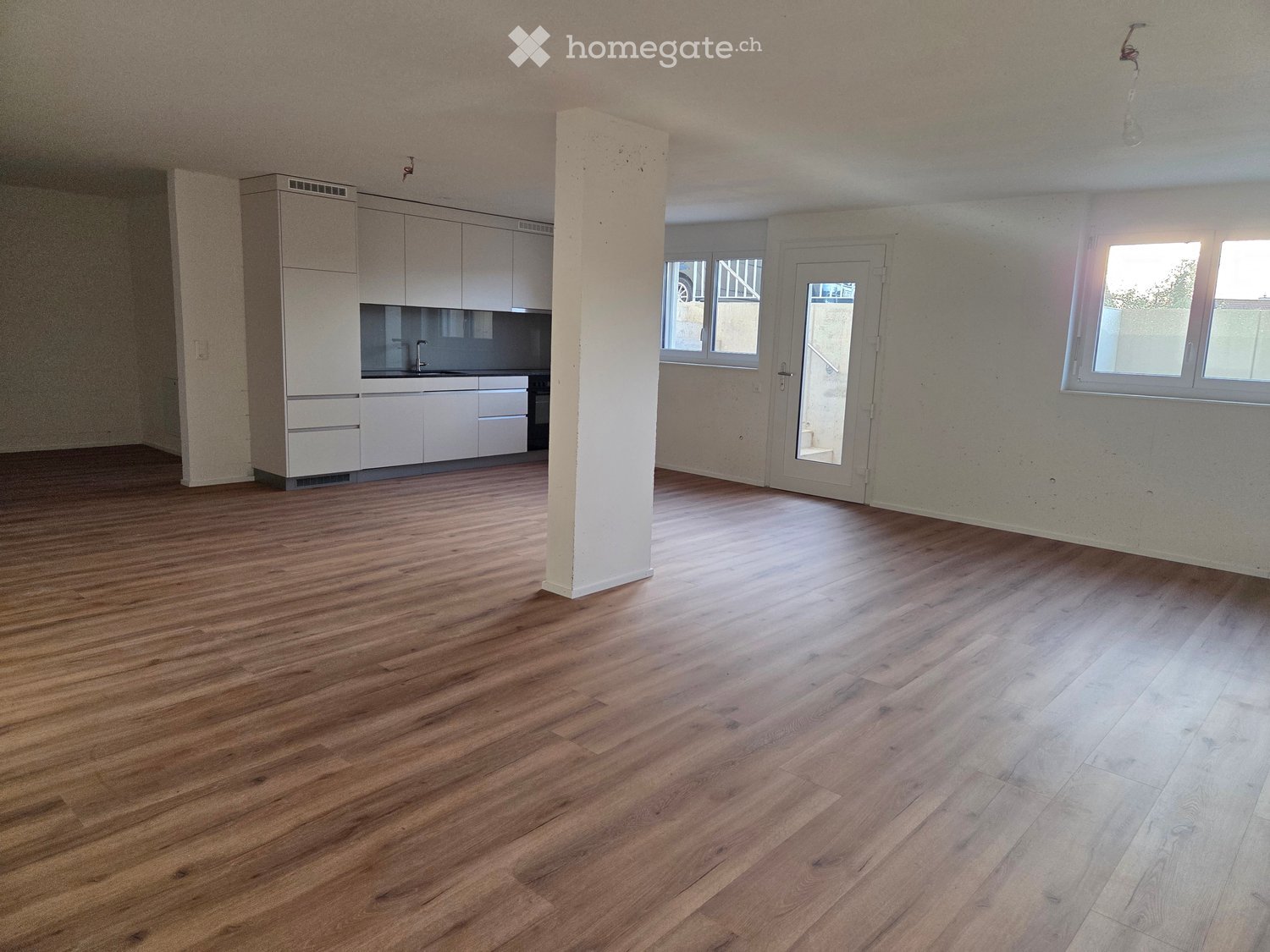 Empty room with wooden flooring, white walls, and a kitchen area with cabinets and a sink visible in the background.