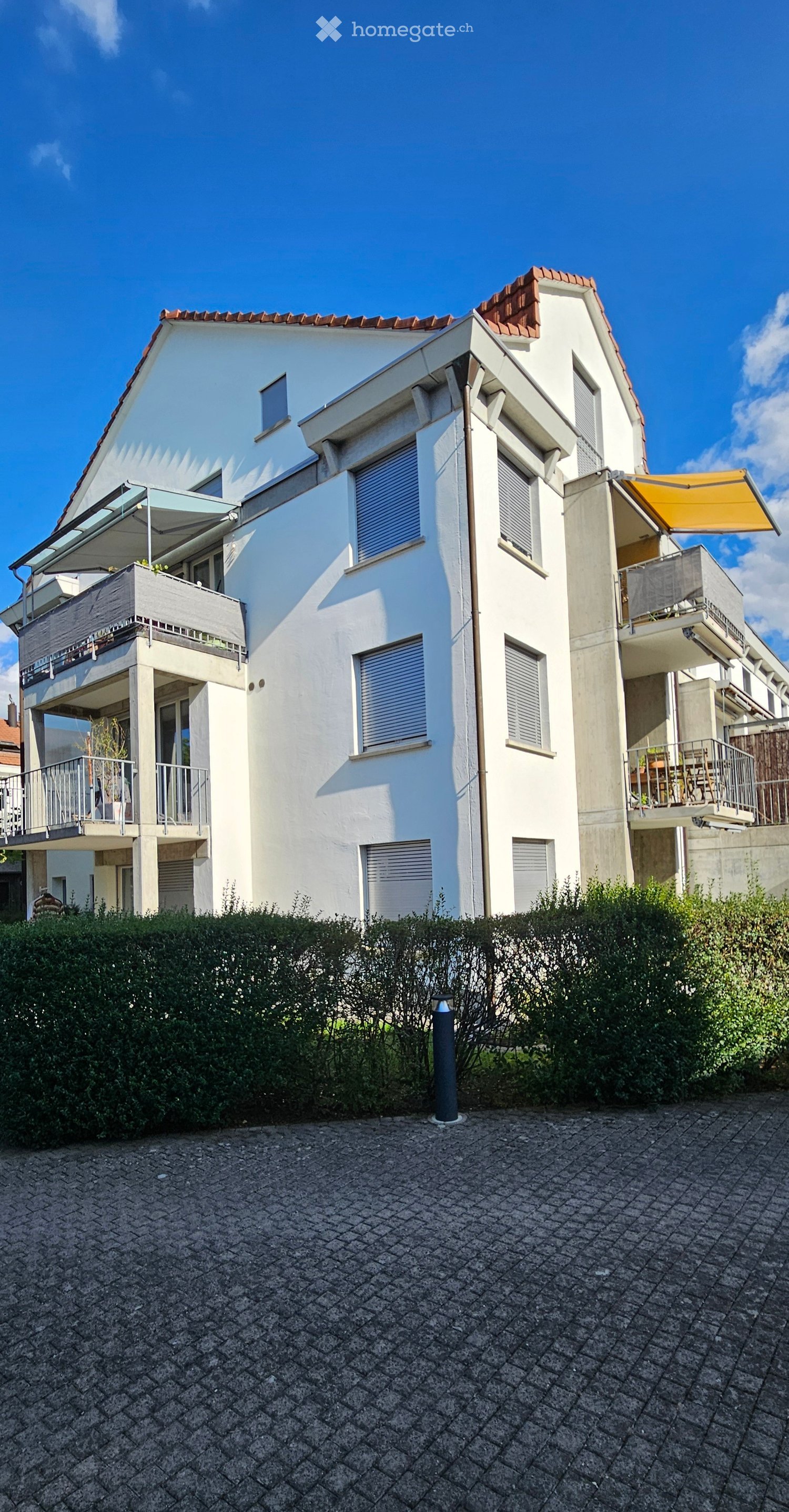 Modern two-story house with white exterior, red-tiled roof, multiple balconies, and a well-maintained hedge in front.