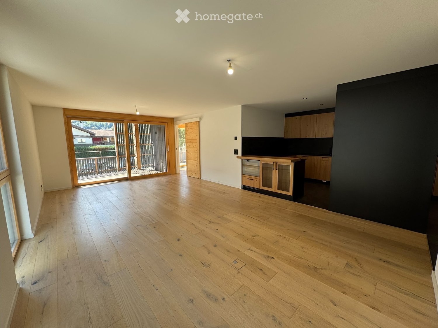 Empty room with wooden flooring, large sliding glass doors leading to a balcony, and a small kitchen area with cabinets and a dishwasher.