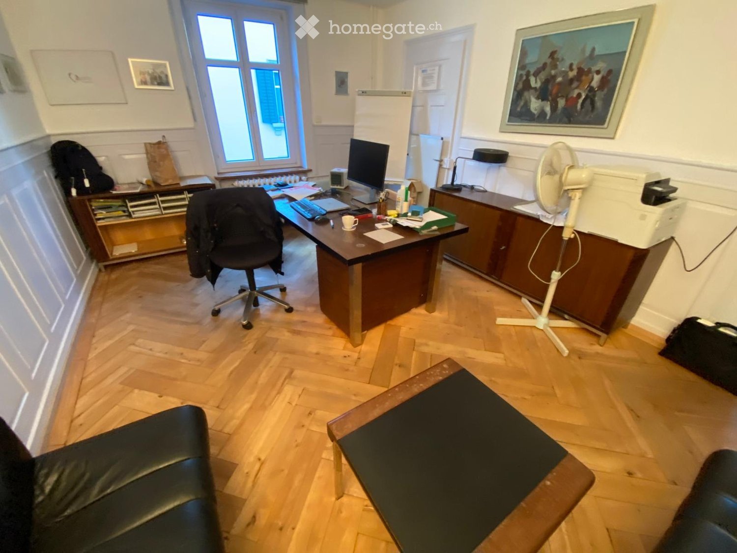 Office space with wooden flooring, large window, desk with computer, office chair, fan, sofa, and a small table.
