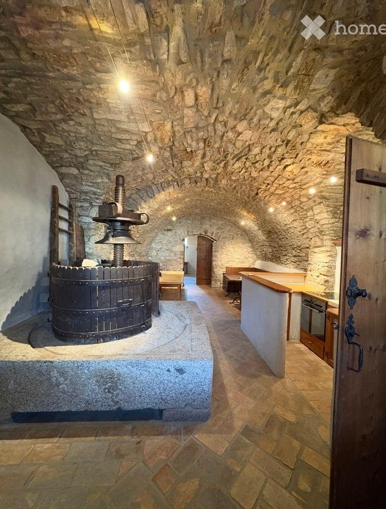 Stone-walled room with a large wooden wine press, a counter, a stool, and an oven.