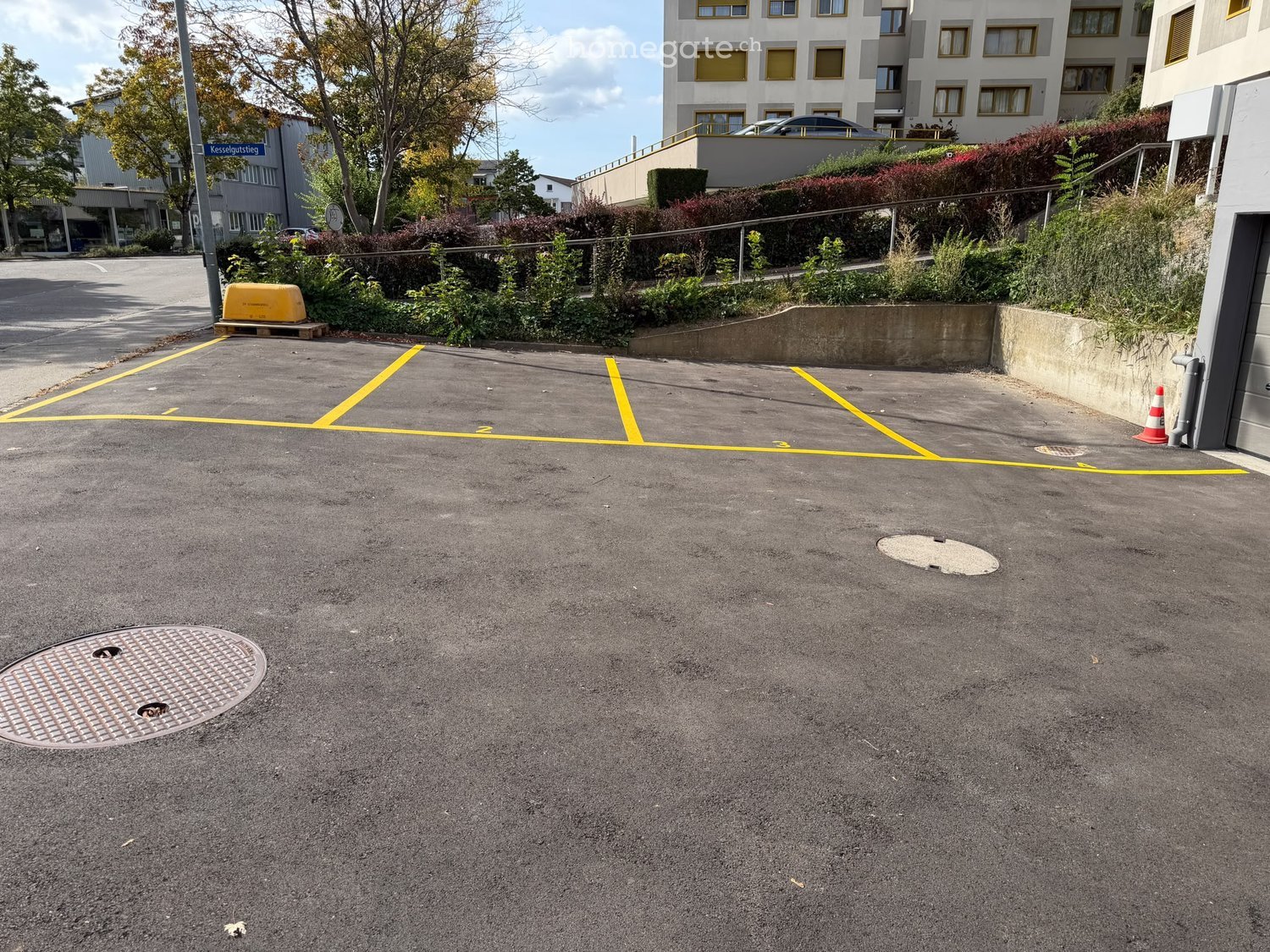 {"type": "Parking place", "objects": [], "description": "Empty parking lot with marked spaces, manhole covers, and a traffic cone. The area is bordered by greenery and a multi-story building in the background.", "localized_descriptions": {"EN": "Empty par