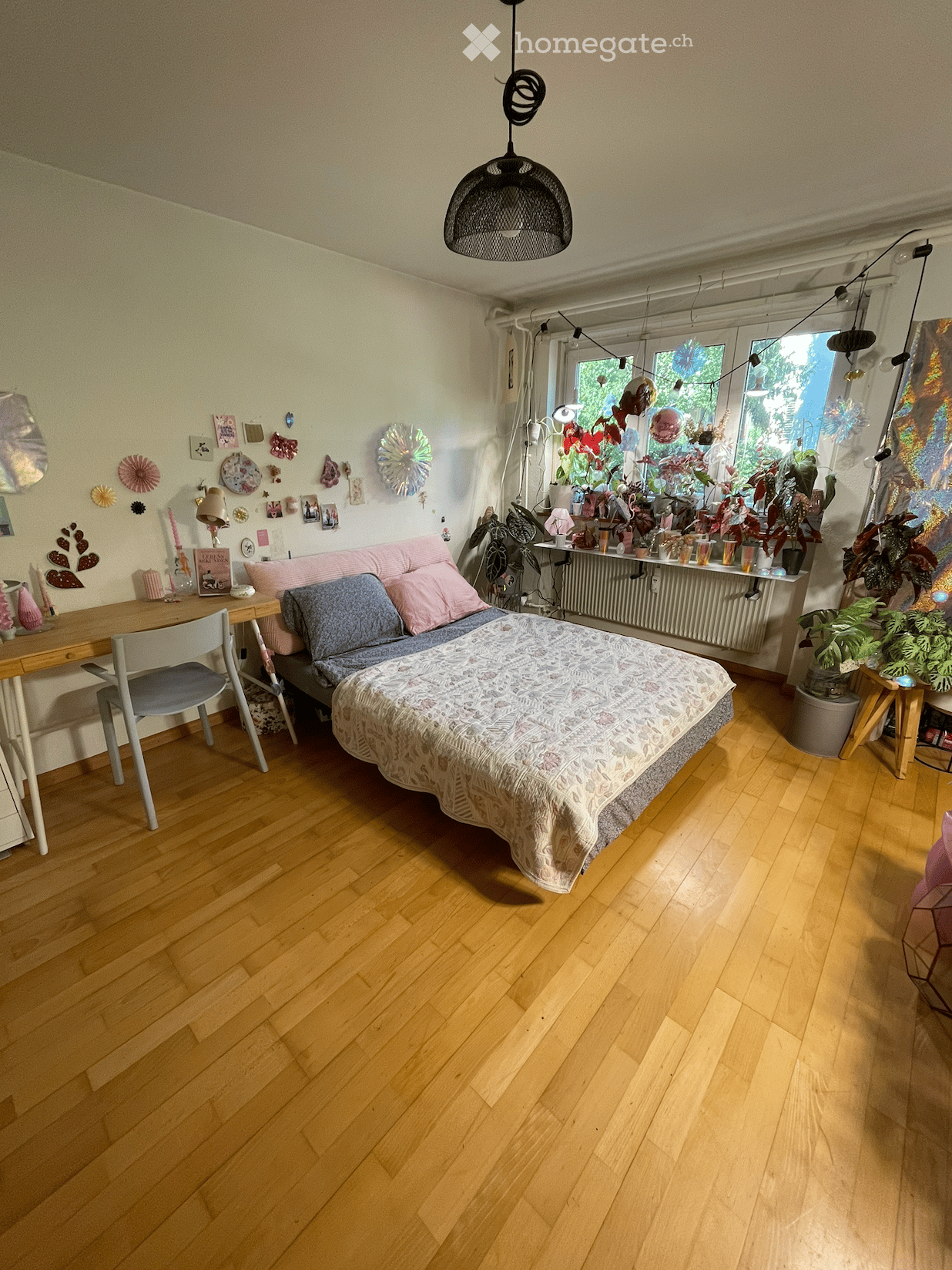 Cozy bedroom with wooden flooring, a bed with patterned bedding, a small sofa, a desk with a chair, and various decorative items and plants on the windowsill and walls.