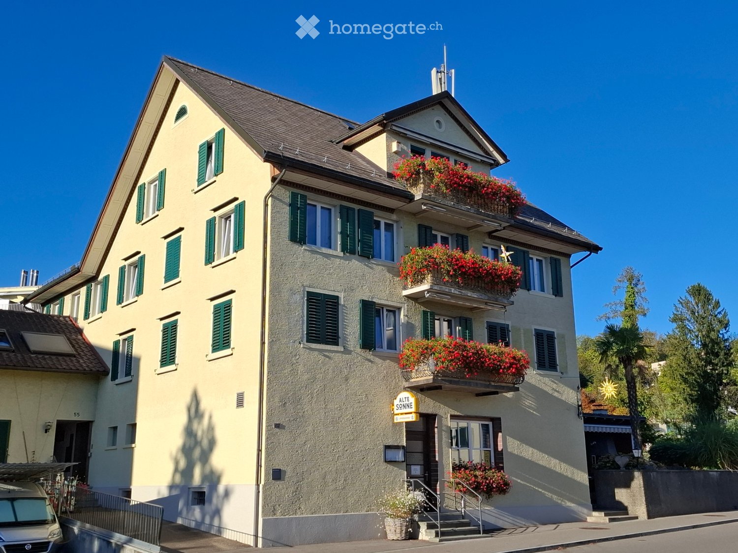 Two-story residential building with green shutters, balconies adorned with red flowers, and a sign reading "ALTE SONNE".