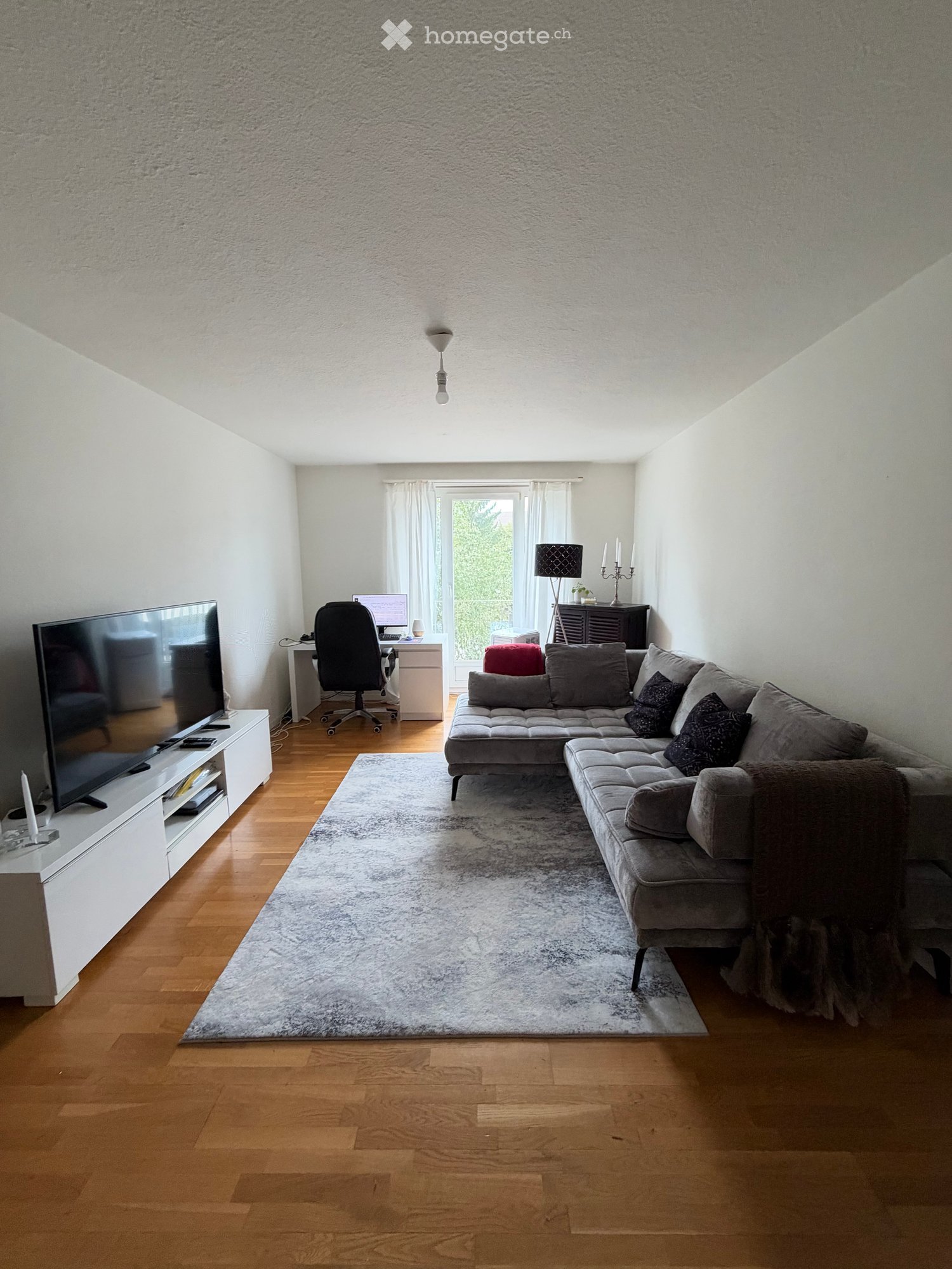 Modern living room with a grey sectional sofa, coffee table, large screen, and a small desk area with a chair and computer. The room features wooden flooring, a large area rug, and a window with sheer curtains.