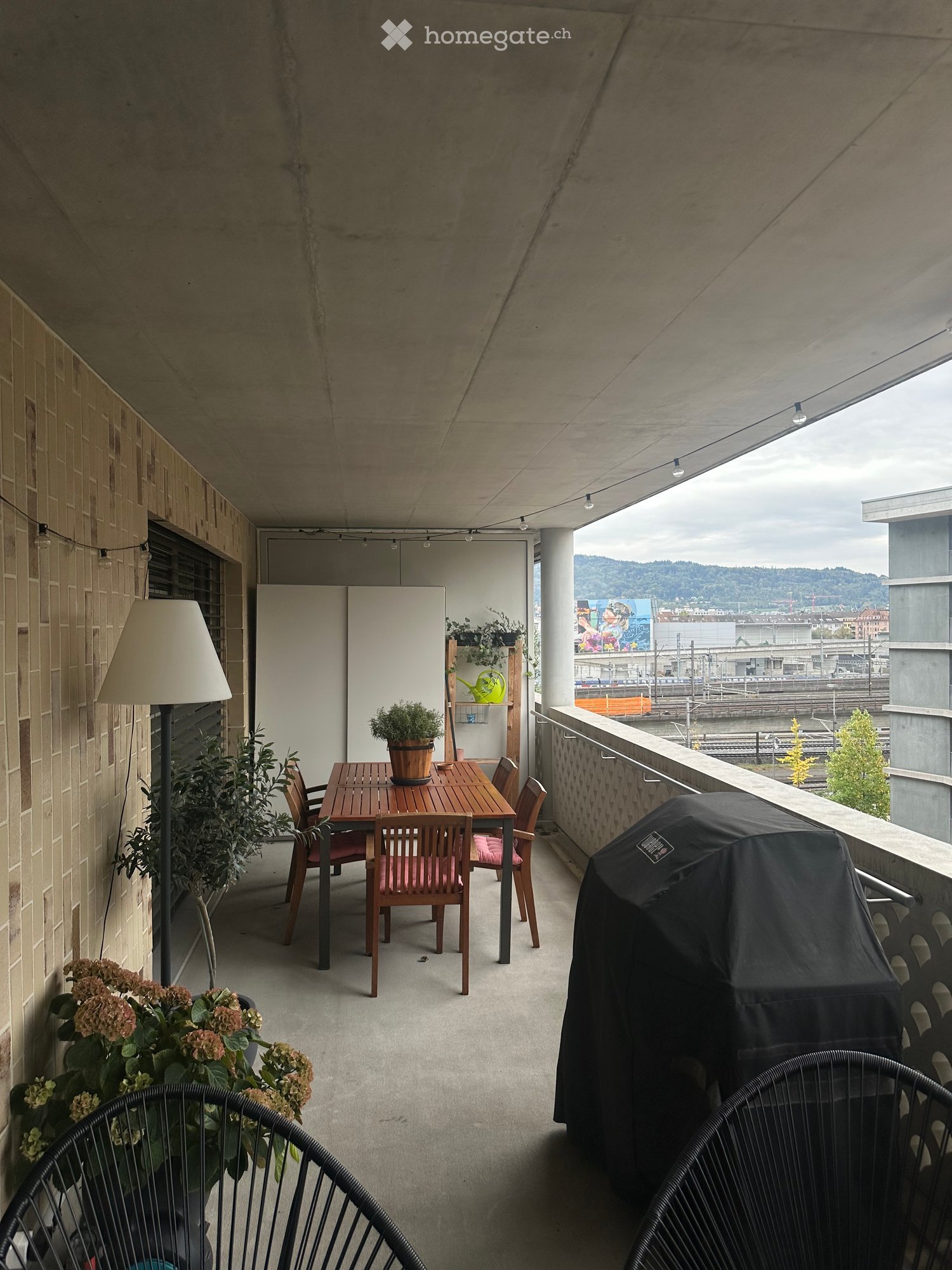 Balcony with a wooden table, chairs, potted plants, a floor lamp, and a covered barbecue, overlooking a cityscape.