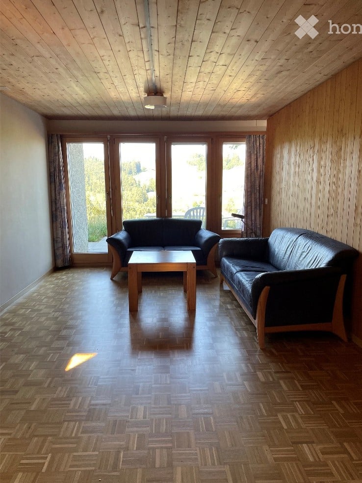Bright living room with wooden parquet flooring, two black leather sofas, a wooden coffee table, large windows with curtains, and a view of the outdoors.