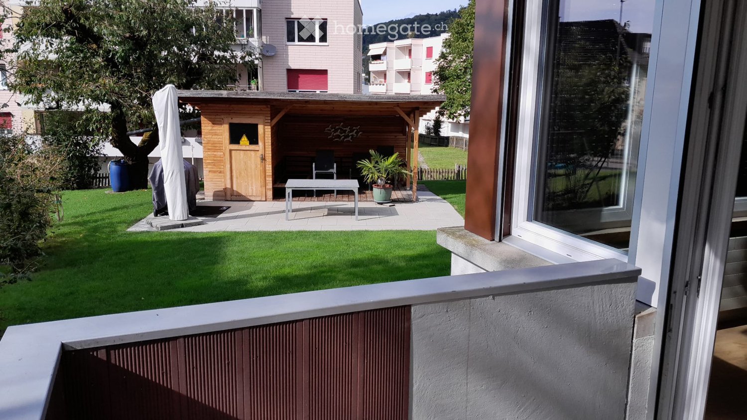 Garden area with a small wooden shelter, a table, chairs, an umbrella, and potted plants. The area is surrounded by a well-maintained lawn and is visible from a balcony.
