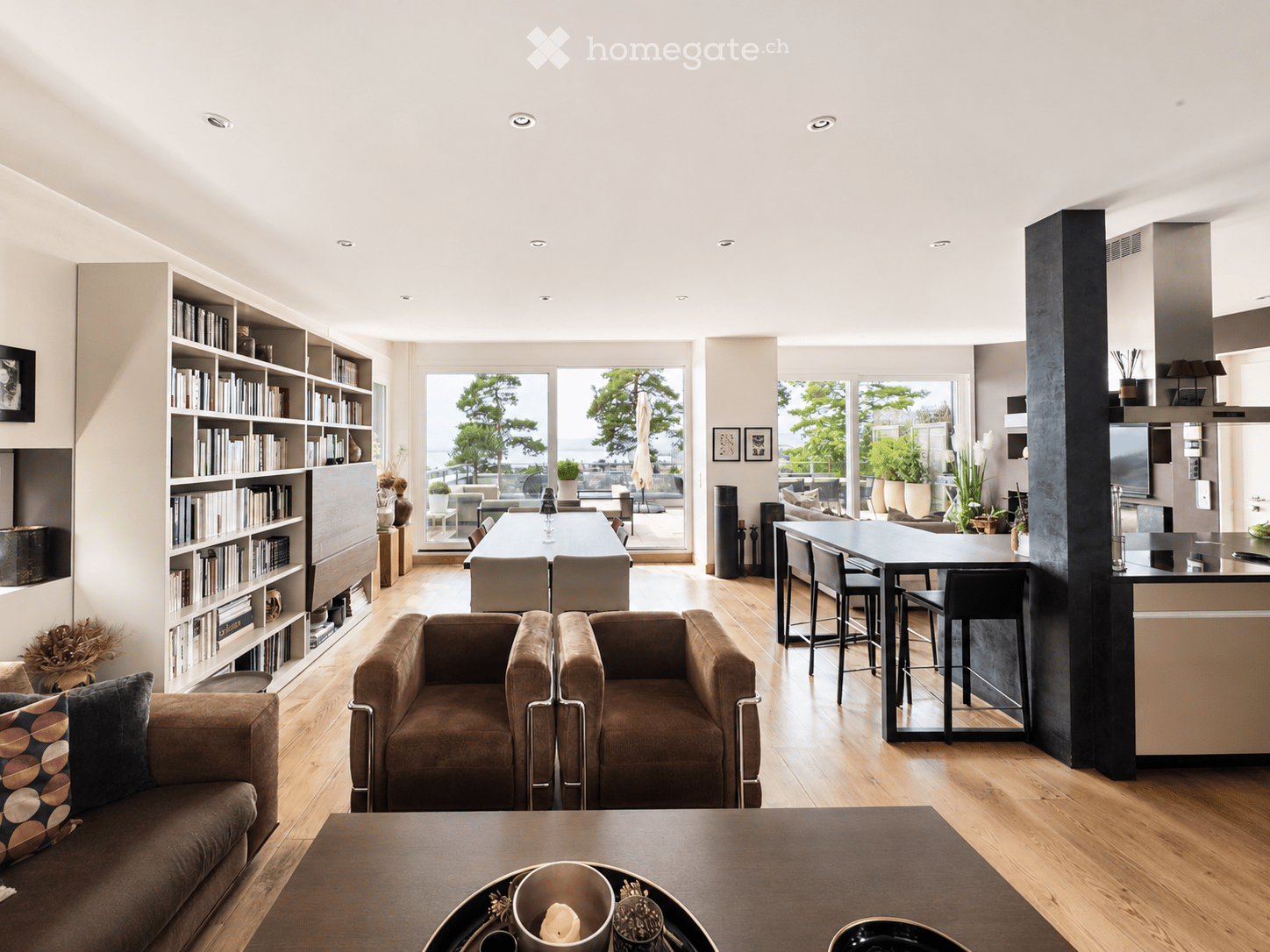 Open-plan living and dining area with wooden flooring, large windows offering a scenic view, a sofa, armchairs, coffee table, dining table, kitchen island, and bar stools.