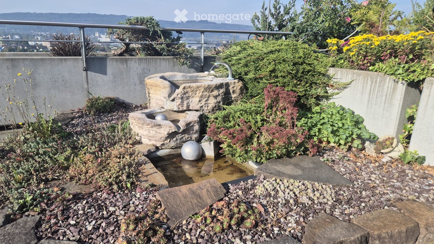 A small, landscaped garden area with a stone water fountain, various plants, and a metal railing overlooking a cityscape.