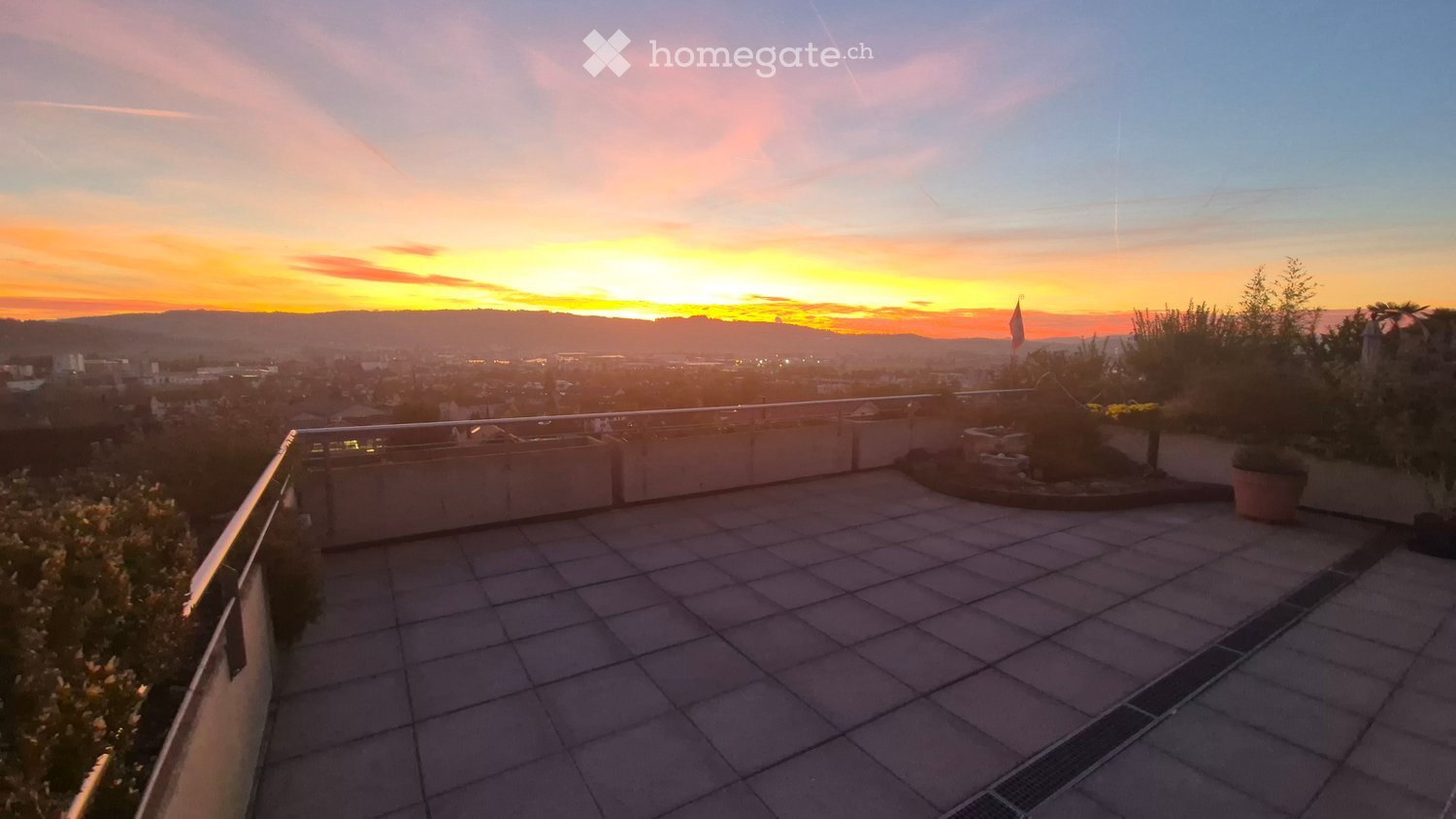 Roof terrace with tiled flooring, metal railing, potted plants, and a panoramic cityscape view at sunset.