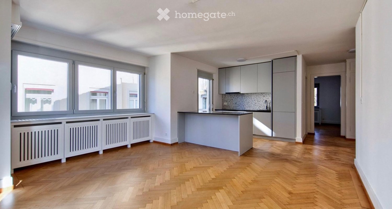 Empty room with wooden herringbone flooring, large windows, radiator covers, and a kitchen area with cabinets and a countertop visible in the background.