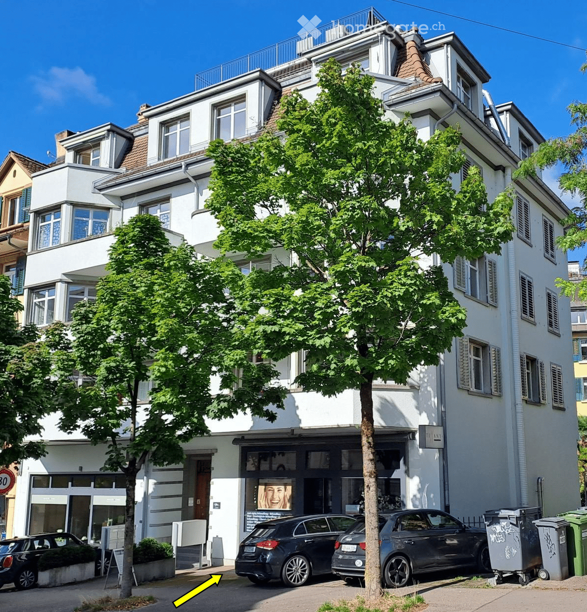 A multi-story residential building with white exterior walls, multiple windows, and a tree in front. There are several parked cars in front of the building.