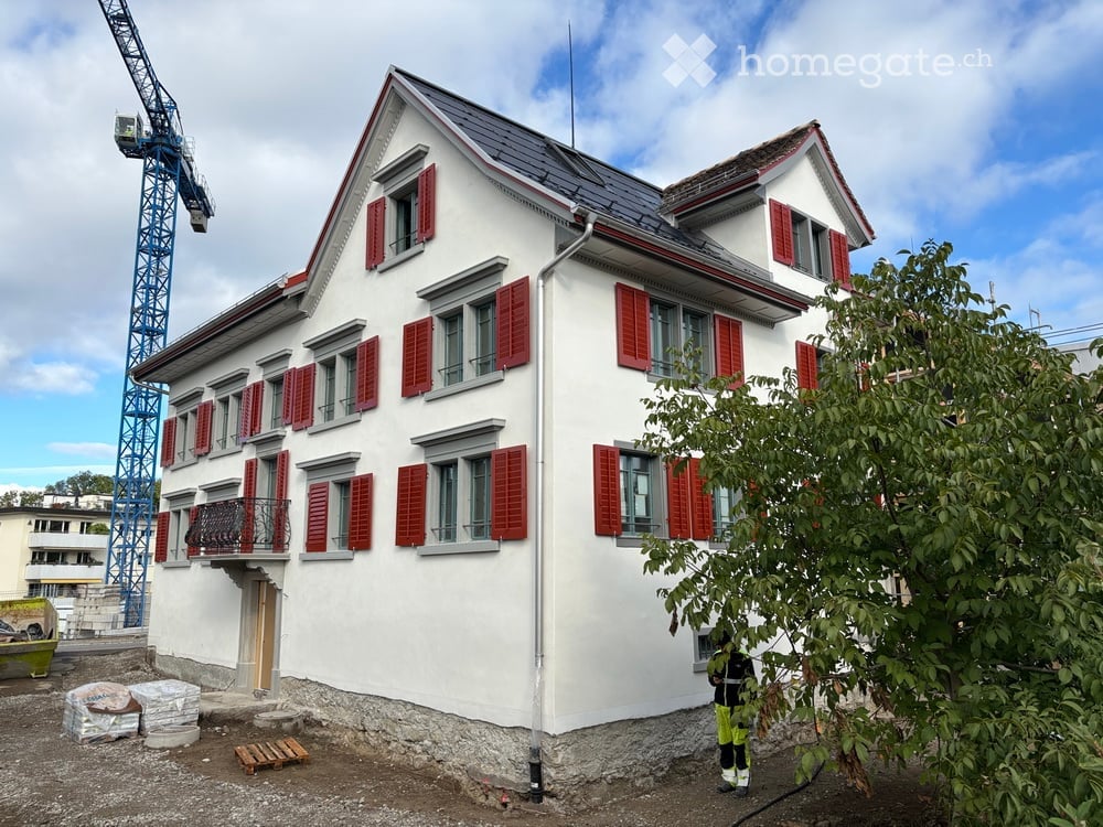 A two-story white house with red shutters and a grey roof, featuring multiple windows and a small balcony on the first floor. The house is surrounded by construction materials and a crane is visible in the background.