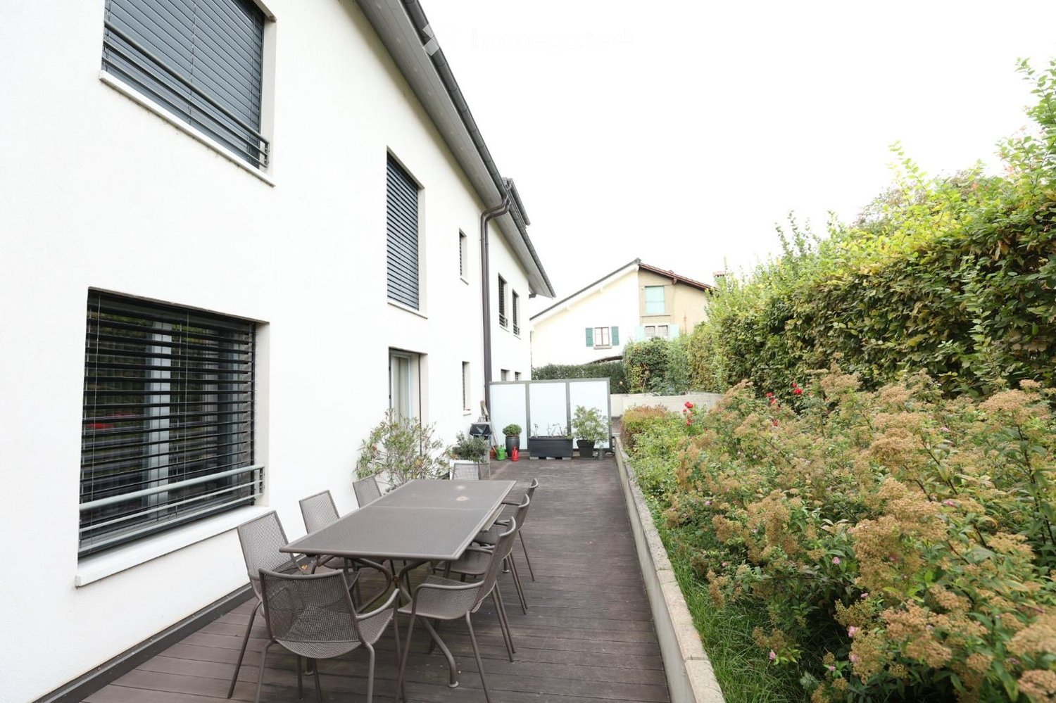 Balcony with wooden decking, grey metal table and chairs, surrounded by greenery and a white building.