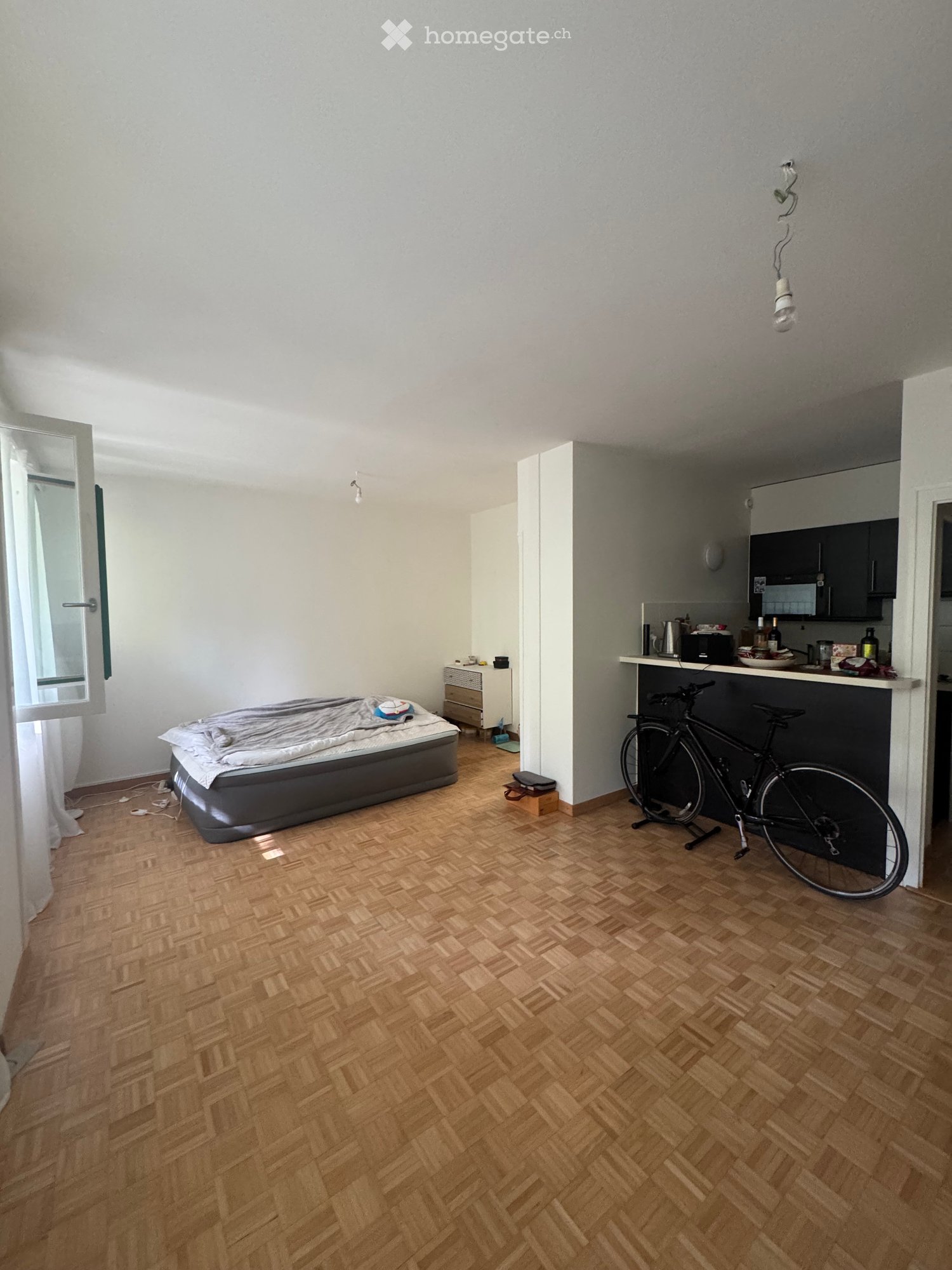 A compact studio apartment with a wooden herringbone floor, a bed with a grey duvet, a small kitchen area with a counter, microwave, fridge, and a bicycle leaning against the wall.