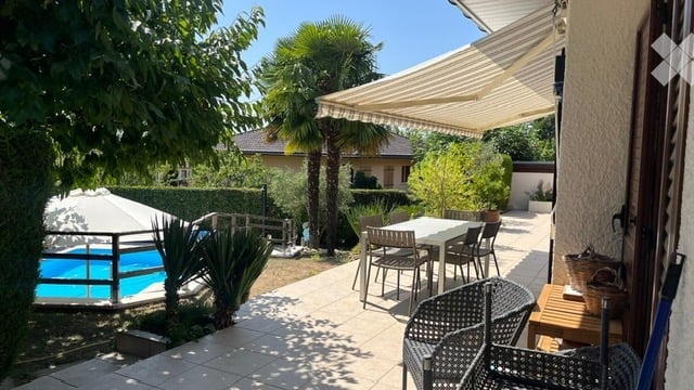 Outdoor patio area with a swimming pool, umbrella, dining table, chairs, and surrounding greenery including palm trees and shrubs.