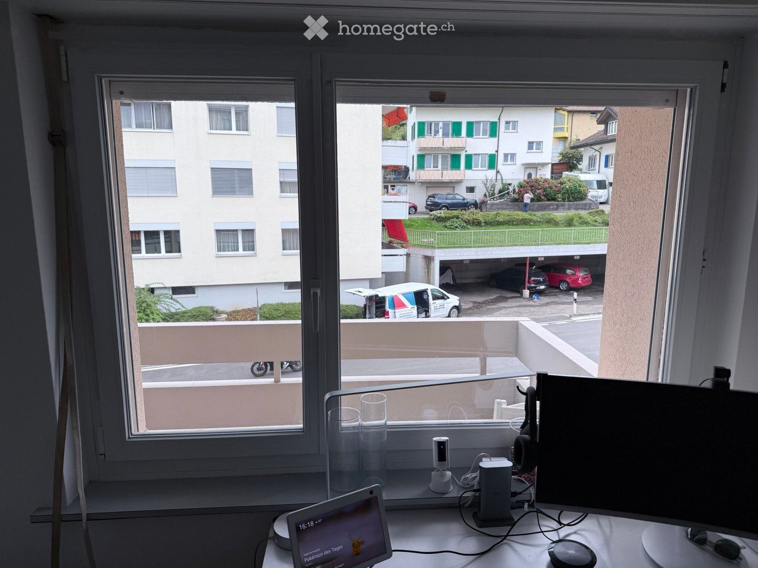 A modern workspace with a laptop, screen, headphones, and laptop stand on a white desk, overlooking a balcony with a view of a residential area and parked cars.