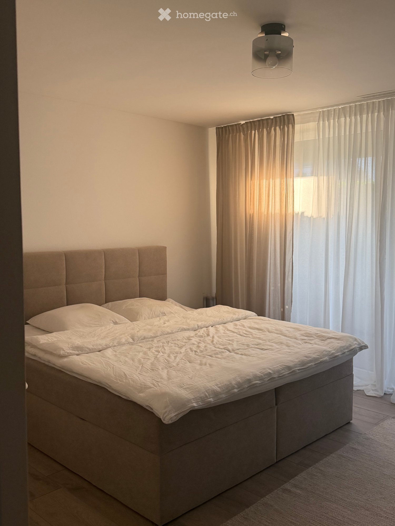 A modern bedroom with a large bed featuring a tufted headboard, white bedding, and light-colored curtains on a window. The room has wooden flooring and a ceiling light fixture.