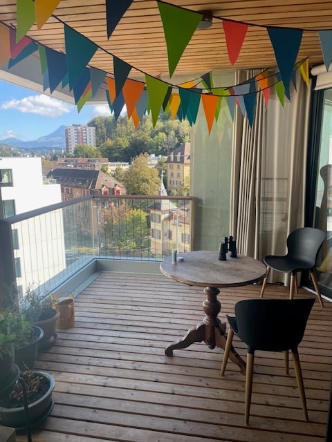 Balcony with wooden decking, small round table, two chairs, potted plants, and colorful triangular flags hanging from the ceiling. Offers a scenic city view with mountains in the background.