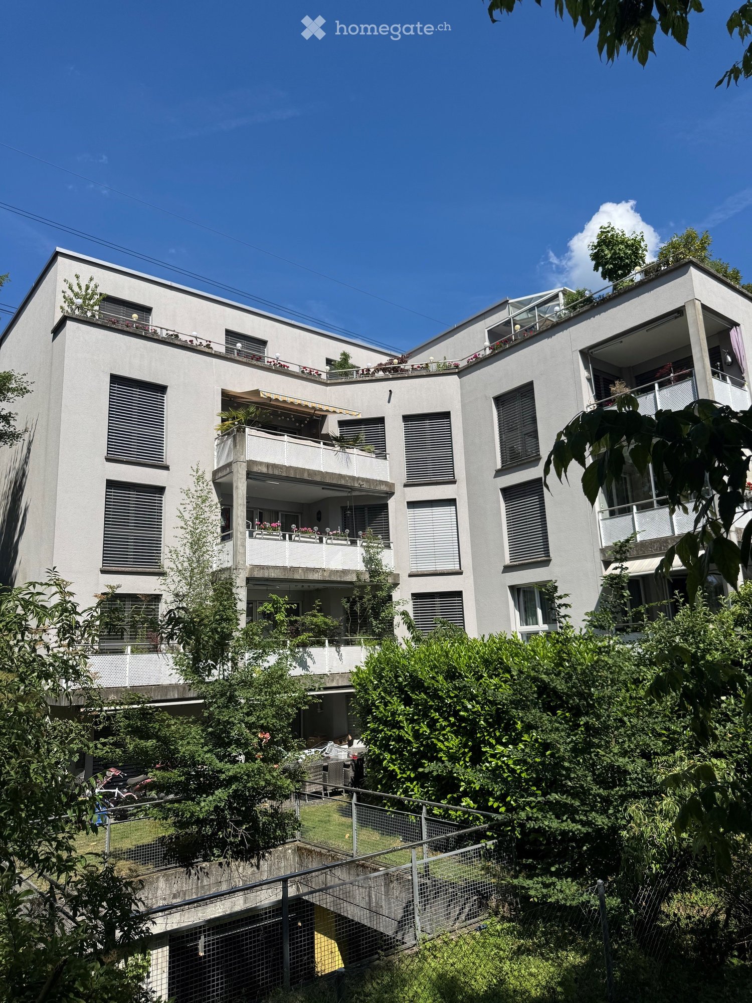 Modern multi-story residential building with balconies, large windows, and greenery on the roof and surrounding area.
