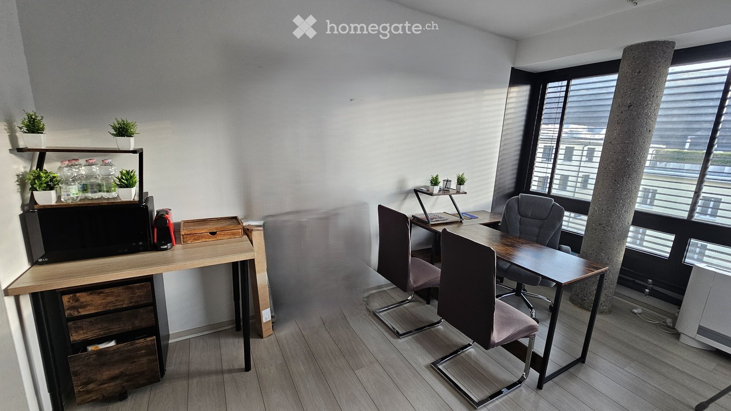 The room has two wooden desks with drawers. A microwave, coffee machine, and potted plants are on the left desk. There are chairs and a wooden desk with a laptop and books on the right.