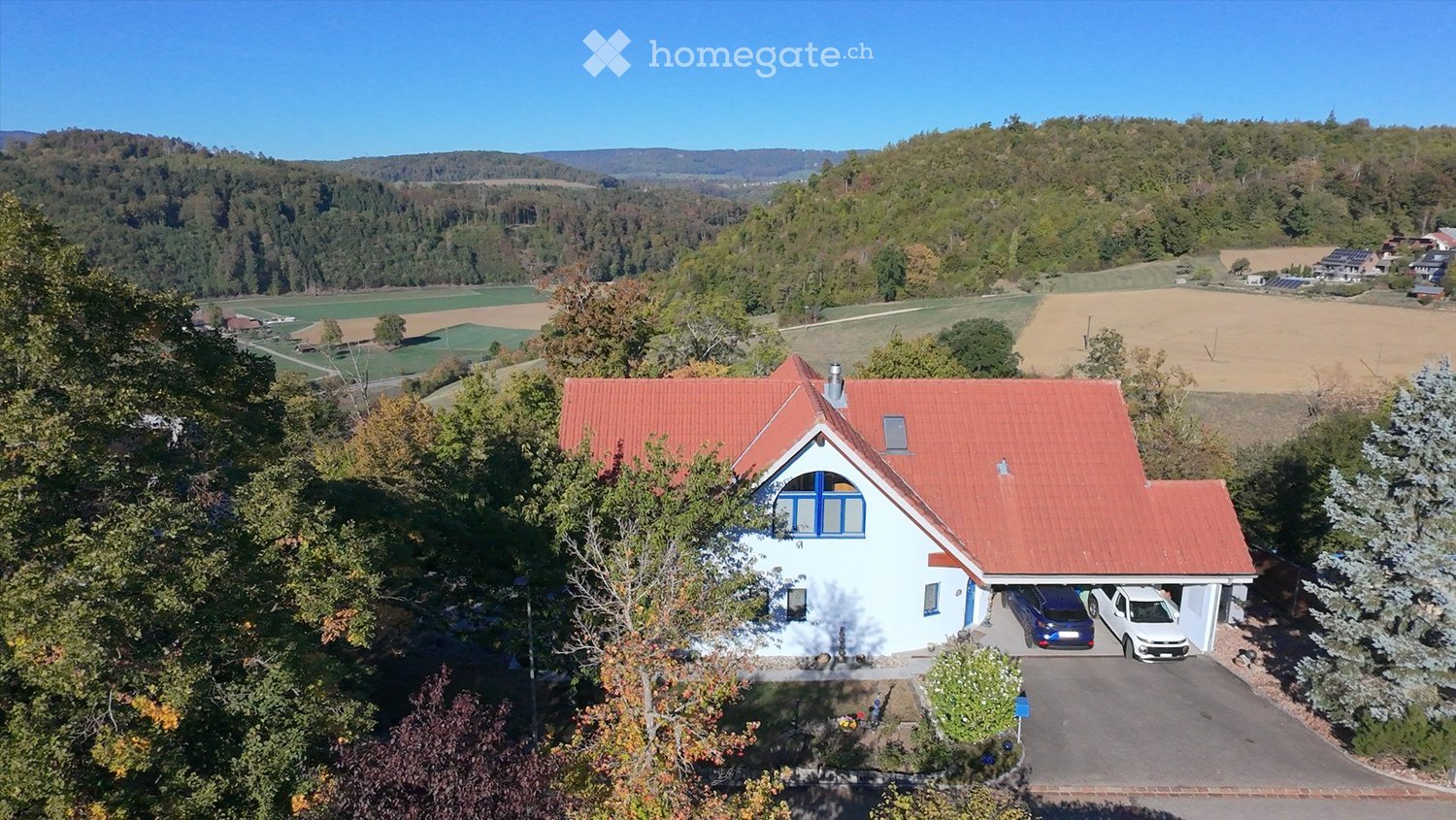 Aerial view of a two-story house with a red-tiled roof, white walls, and blue window frames. The house features a garage with two parked cars and is surrounded by a landscaped garden. The background shows a scenic countryside with fields, trees, and dista