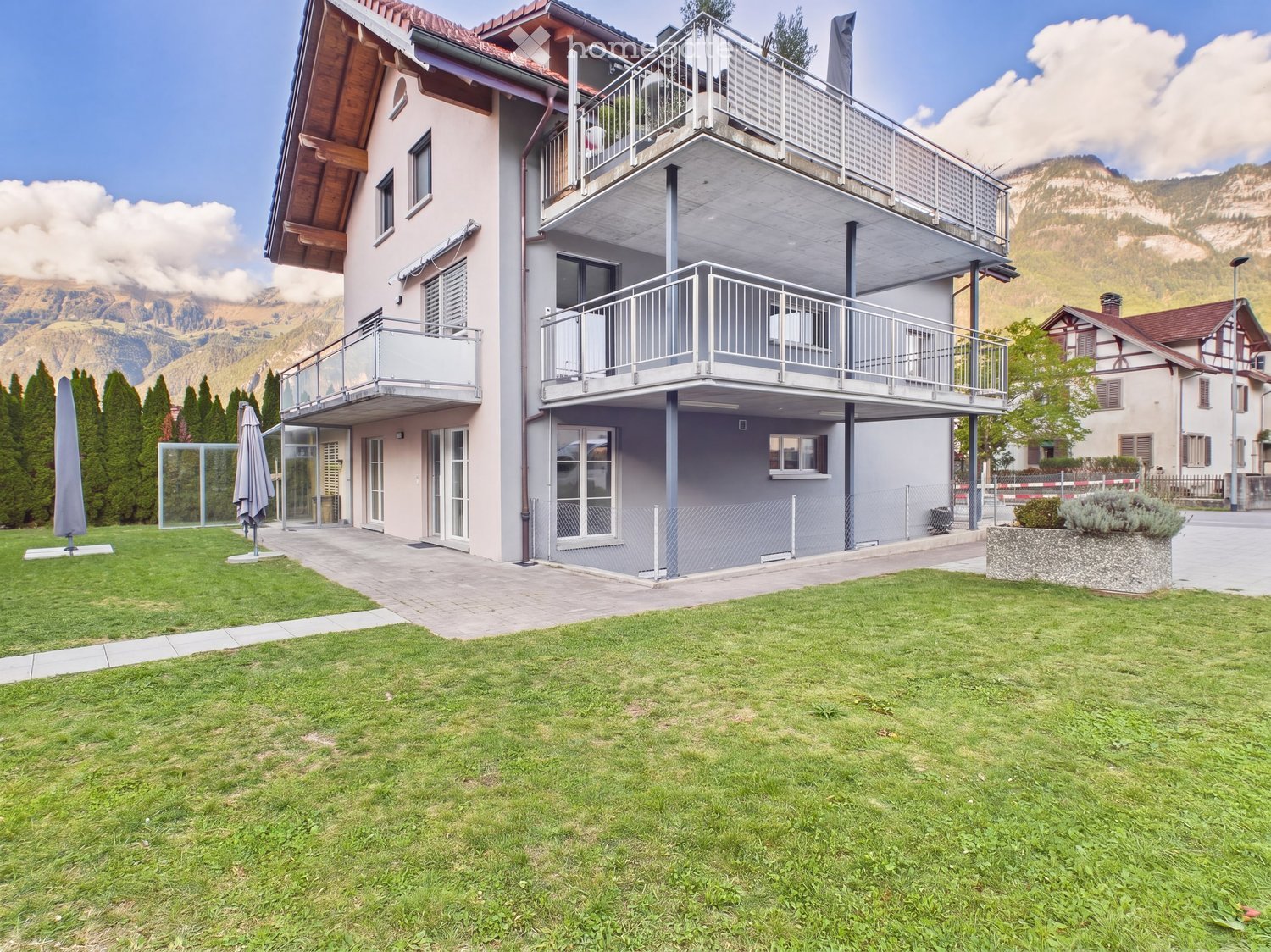 Modern two-story house with balconies, surrounded by a well-maintained lawn and trees, with mountain views in the background.
