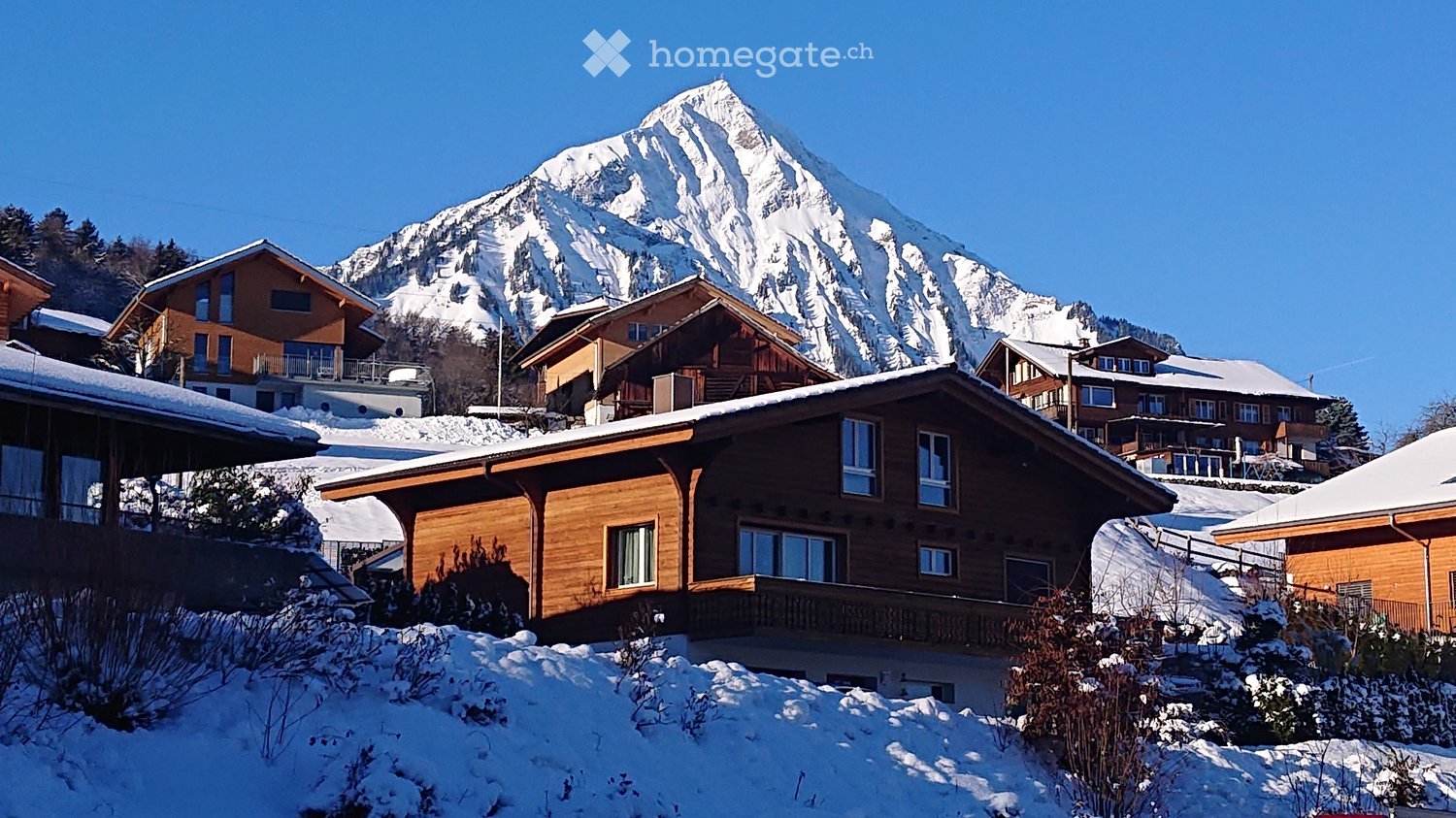A cluster of wooden chalets in a snowy alpine village with a snow-capped mountain in the background.