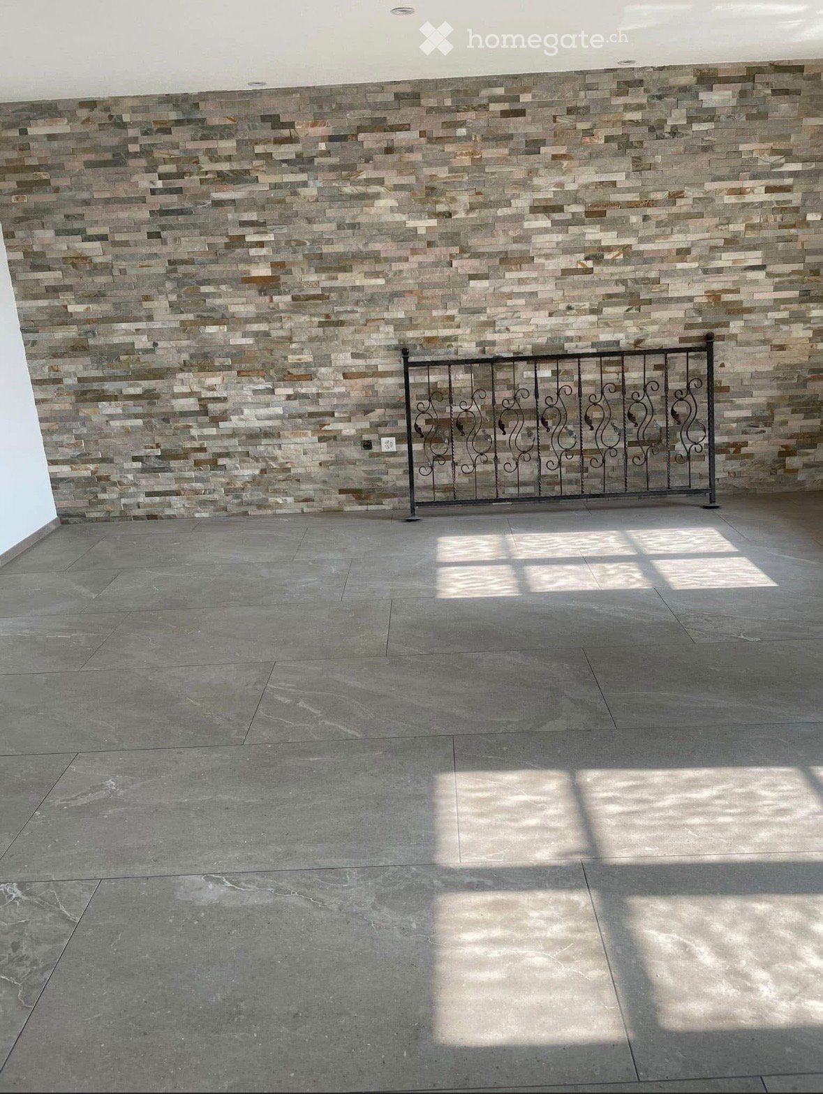 Empty room with a tiled wall and a decorative metal gate, featuring a concrete floor and natural light from an unseen source.