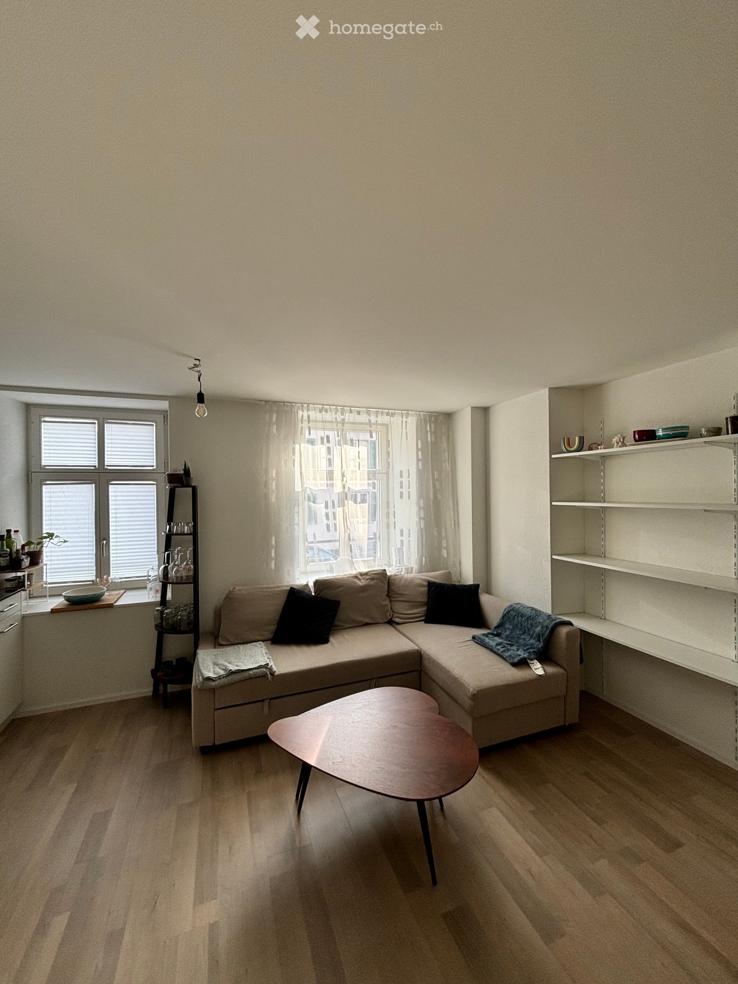 Modern living room with a beige sectional sofa, wooden coffee table, shelves, and large windows with curtains.