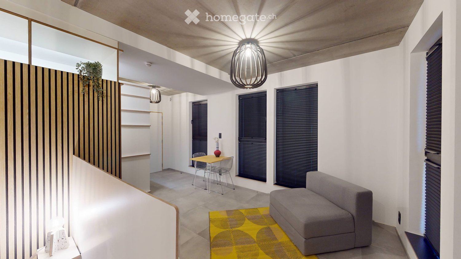 Modern living room with a grey sofa, yellow rug, small dining table with chairs, and decorative vase. Features large windows, pendant lights, and a sleek wooden shelf with a plant.
